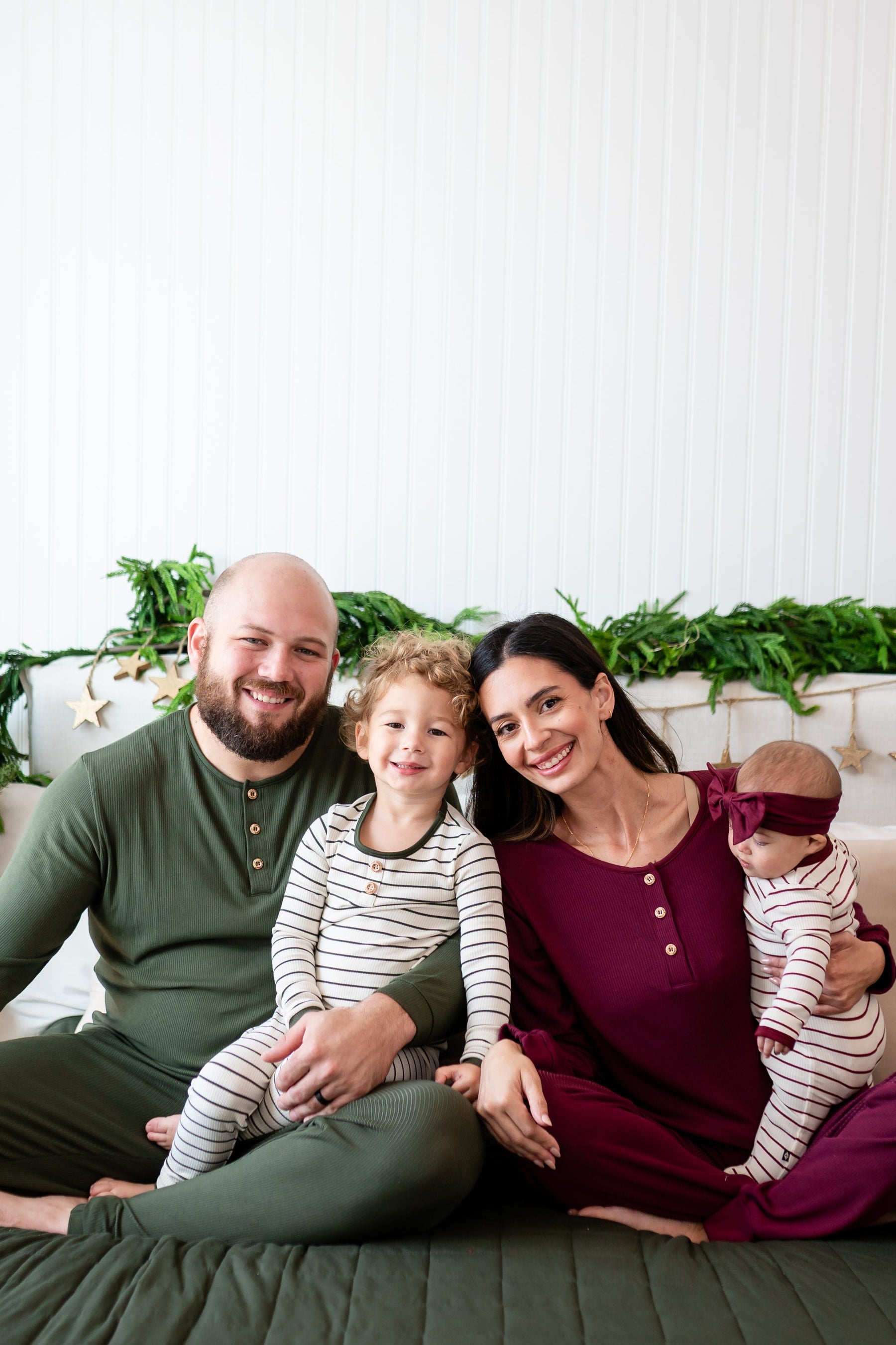 Family of four sitting on a bed wearing the Henley sets in Fir, Fir Strip, Burgundy and Burgundy Stripe