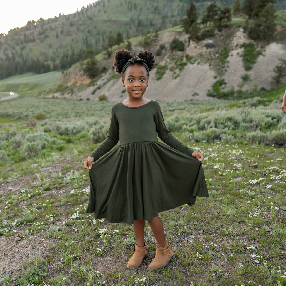 Young girl in a green dress standing in a natural landscape with mountains and greenery.