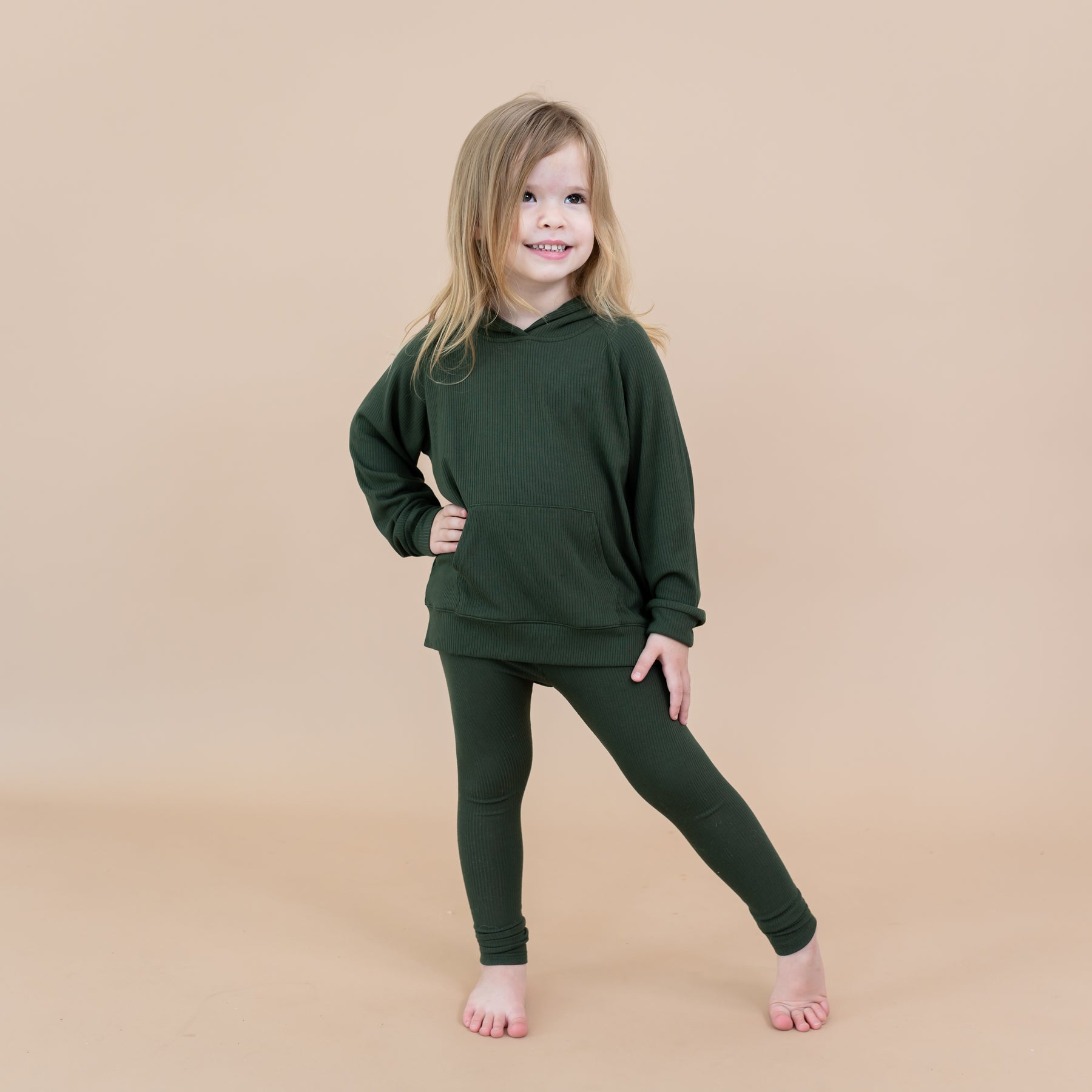 Young girl standing with one leg out to the side wearing the Ribbed Leggings in Fir paired with a matching ribbed hoodie in front of a light beige background