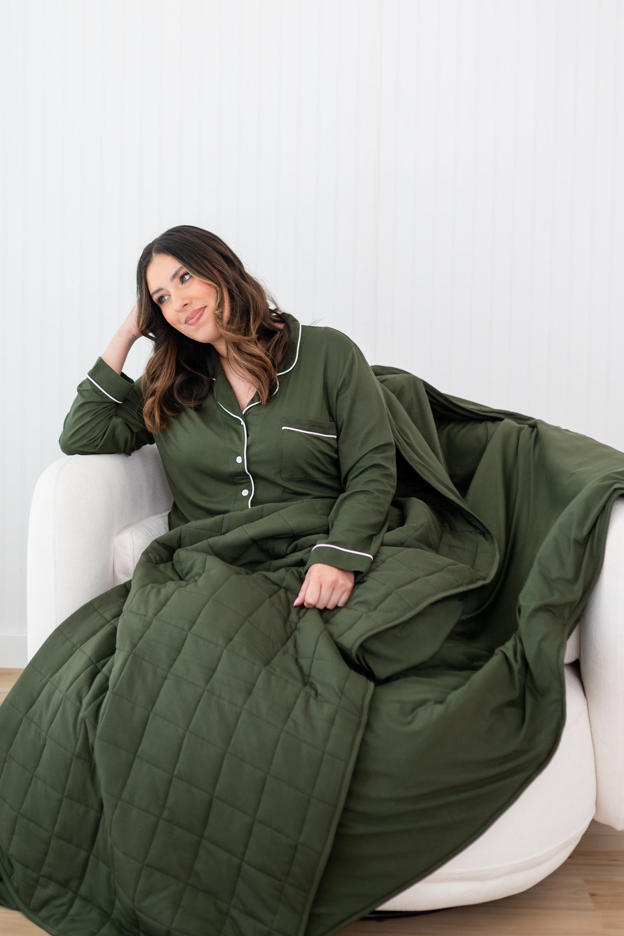 Woman sitting cozily in a white chair with a dark olive green blanket draped over her while wearing matching colored pajamas