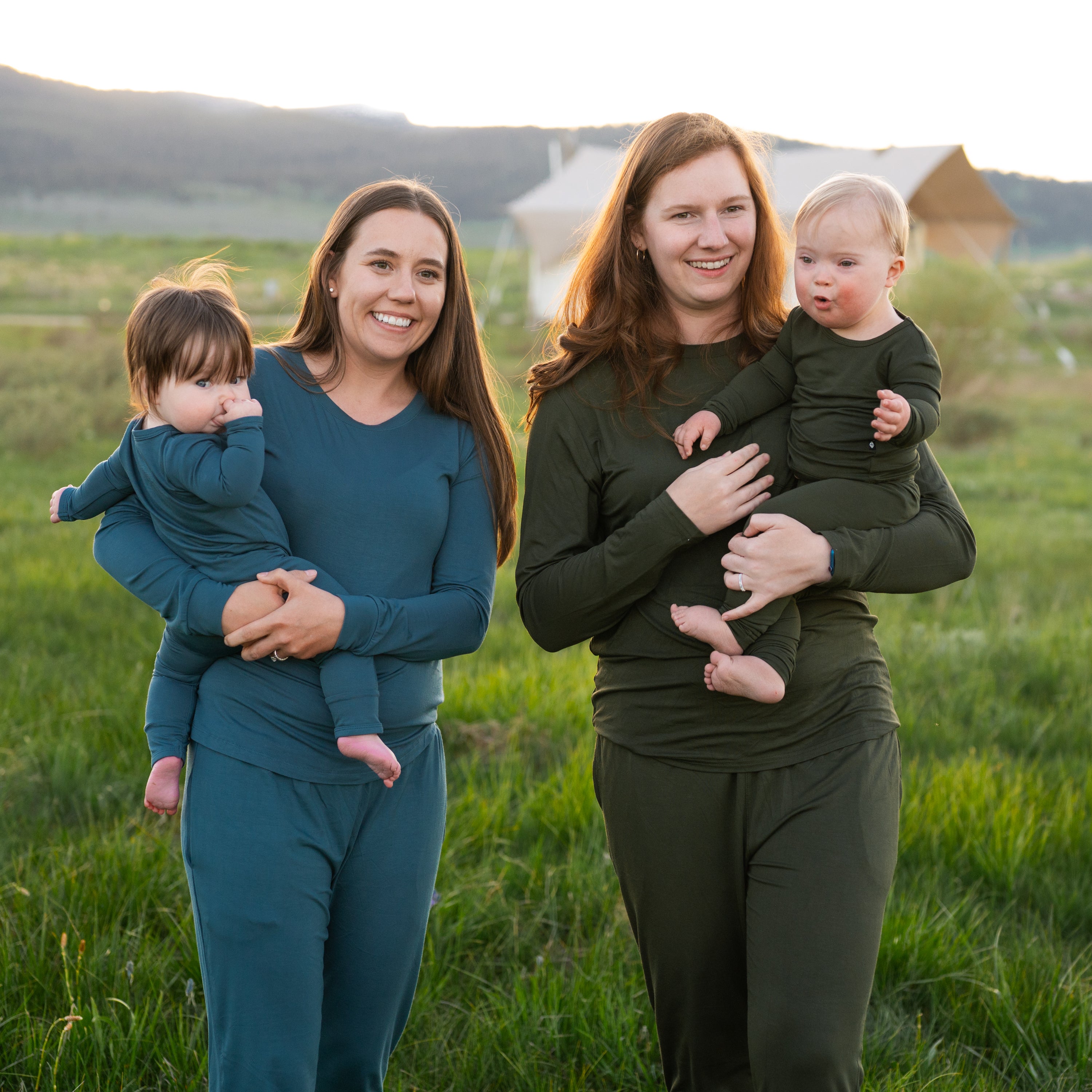 Two women standing in a grassy field, each holding a baby, wearing matching outfits.