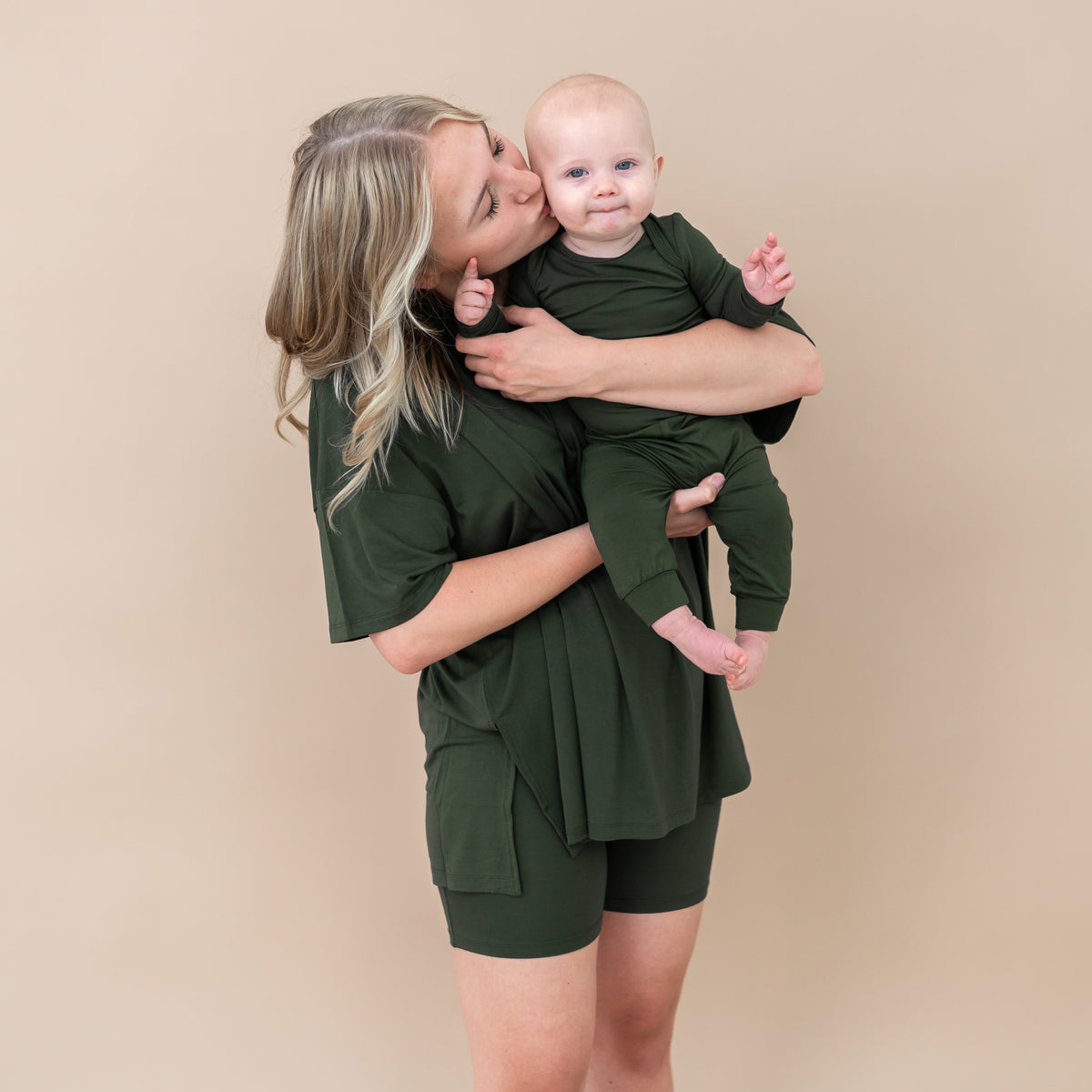 Woman holding a baby both wearing green outfits against a beige background