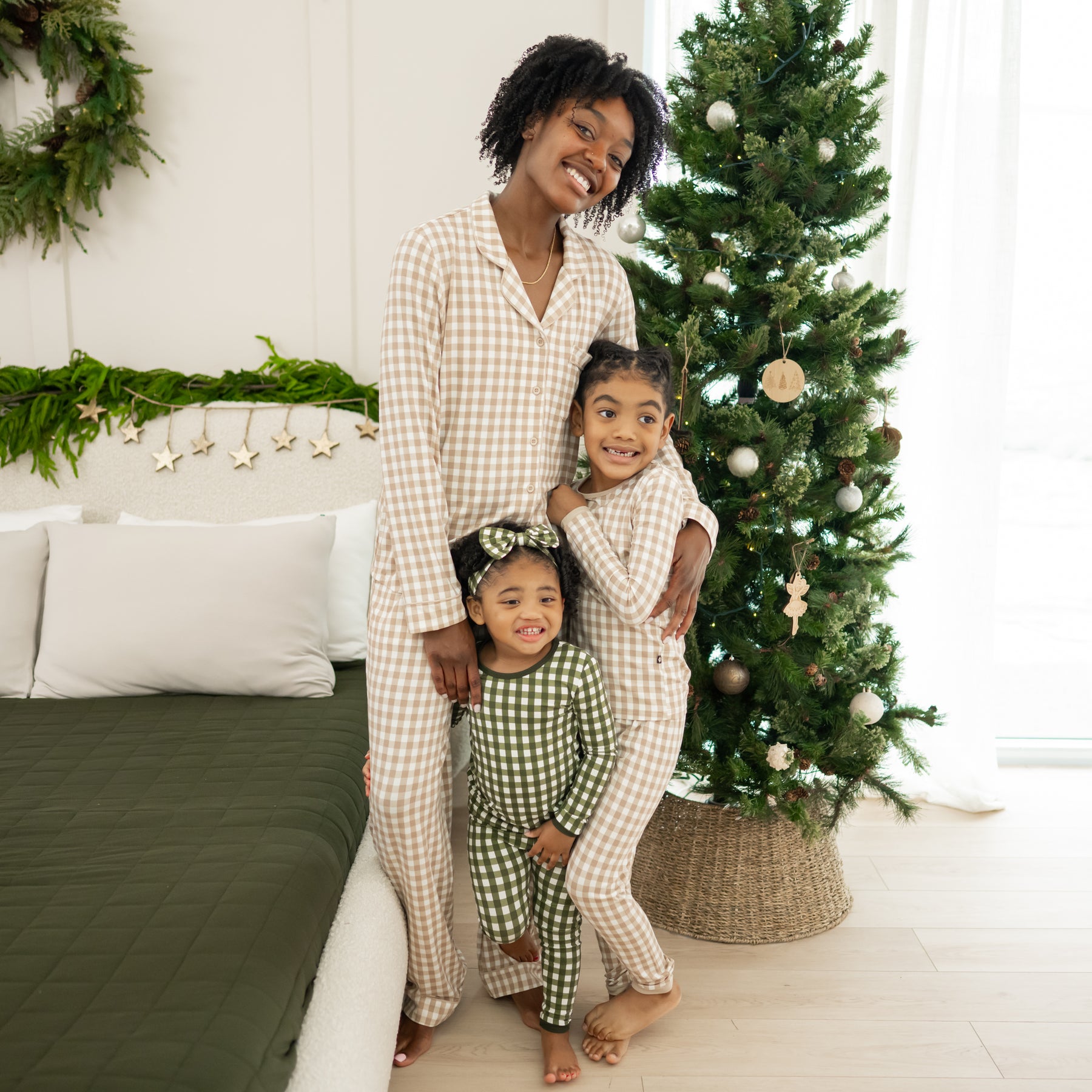 Matching family wearing long sleeve pajamas standing beside a bed in front of a Christmas tree. Mother wearing the Long-Sleeved Women's Pajama Set in Gingham Bisque matching her oldest daughter. Youngest is wearing the Fir gingham toddler pajama set