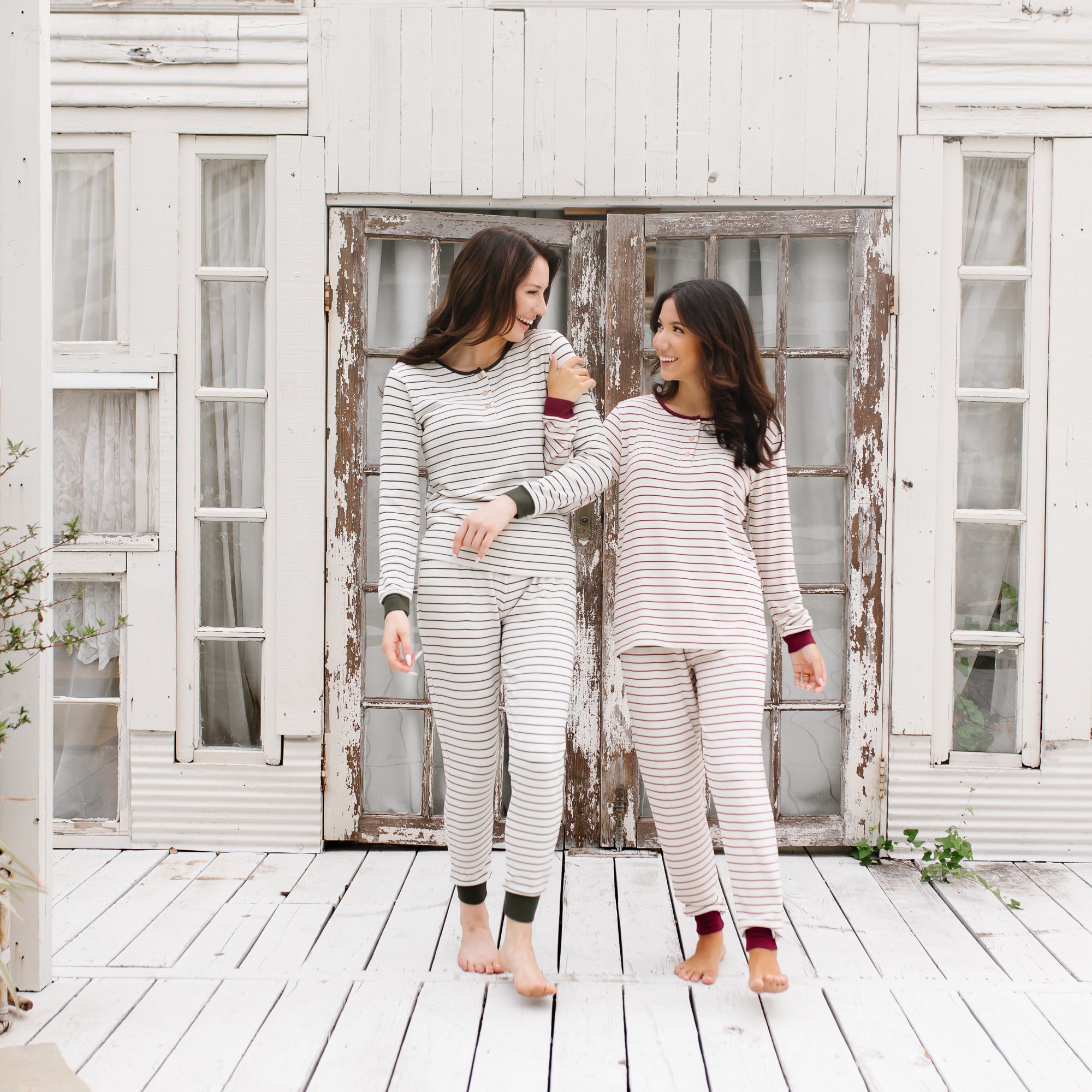 Two females walking arm in arm on a white wooden deck in front of a white wooden house wearing the Women’s Ribbed Henley Set in Fir Stripe and Burgundy Stripe