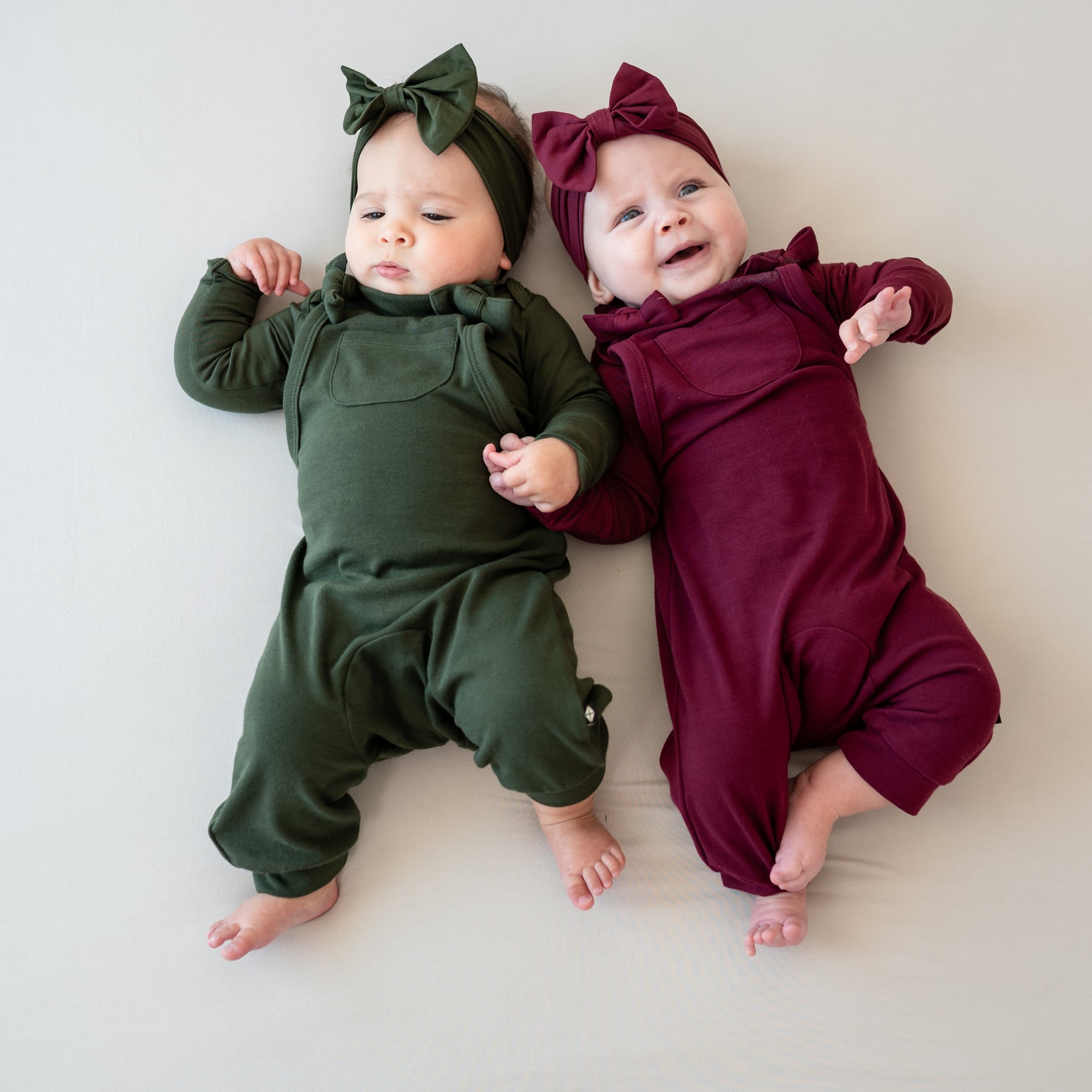 Two infant girls laying side by side wearing the Bamboo Jersey Overalls in Burgundy and Fir and matching long sleeve bodysuits and Bow Headbands