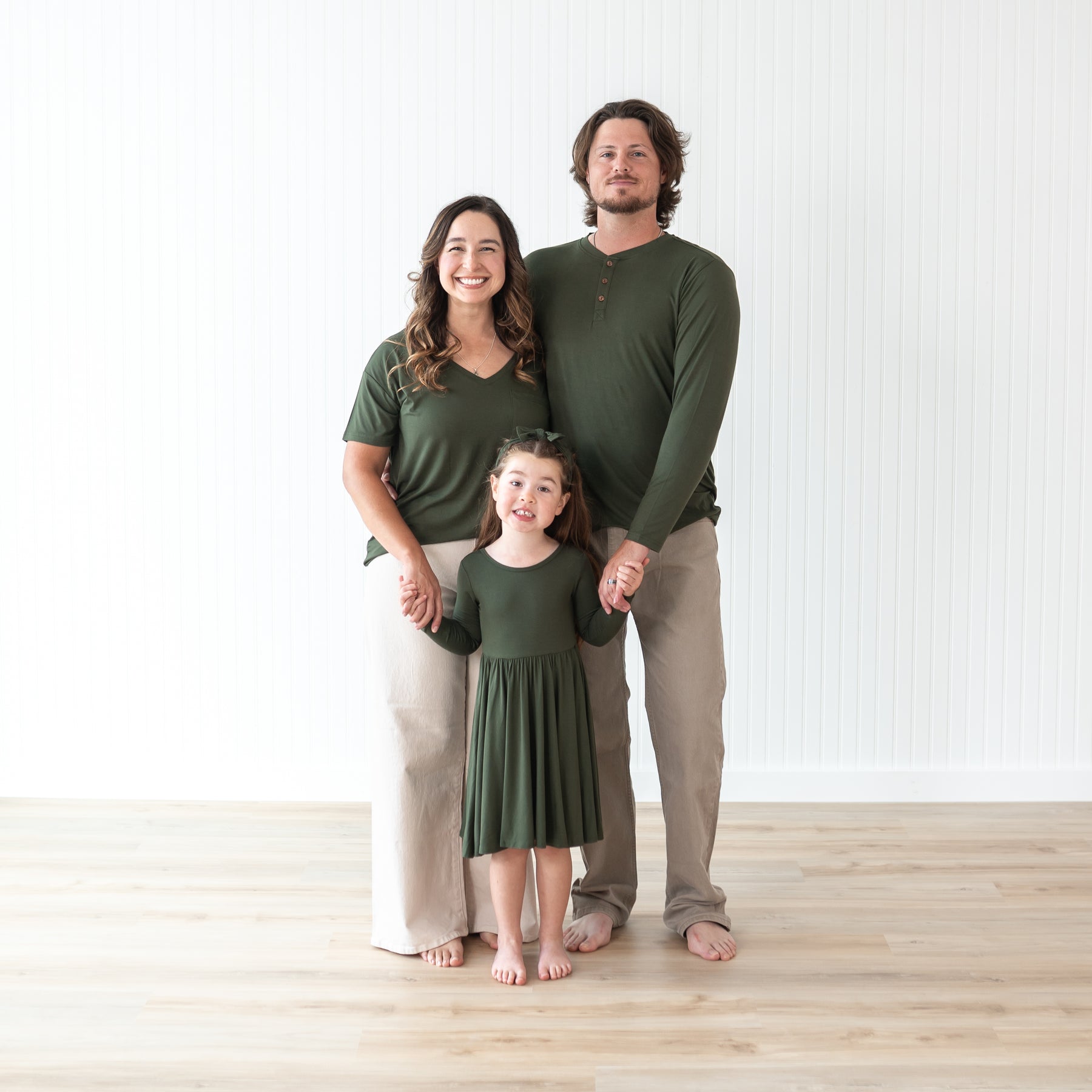 Family of three wearing matching green bamboo outfits standing on a light wooden floor.