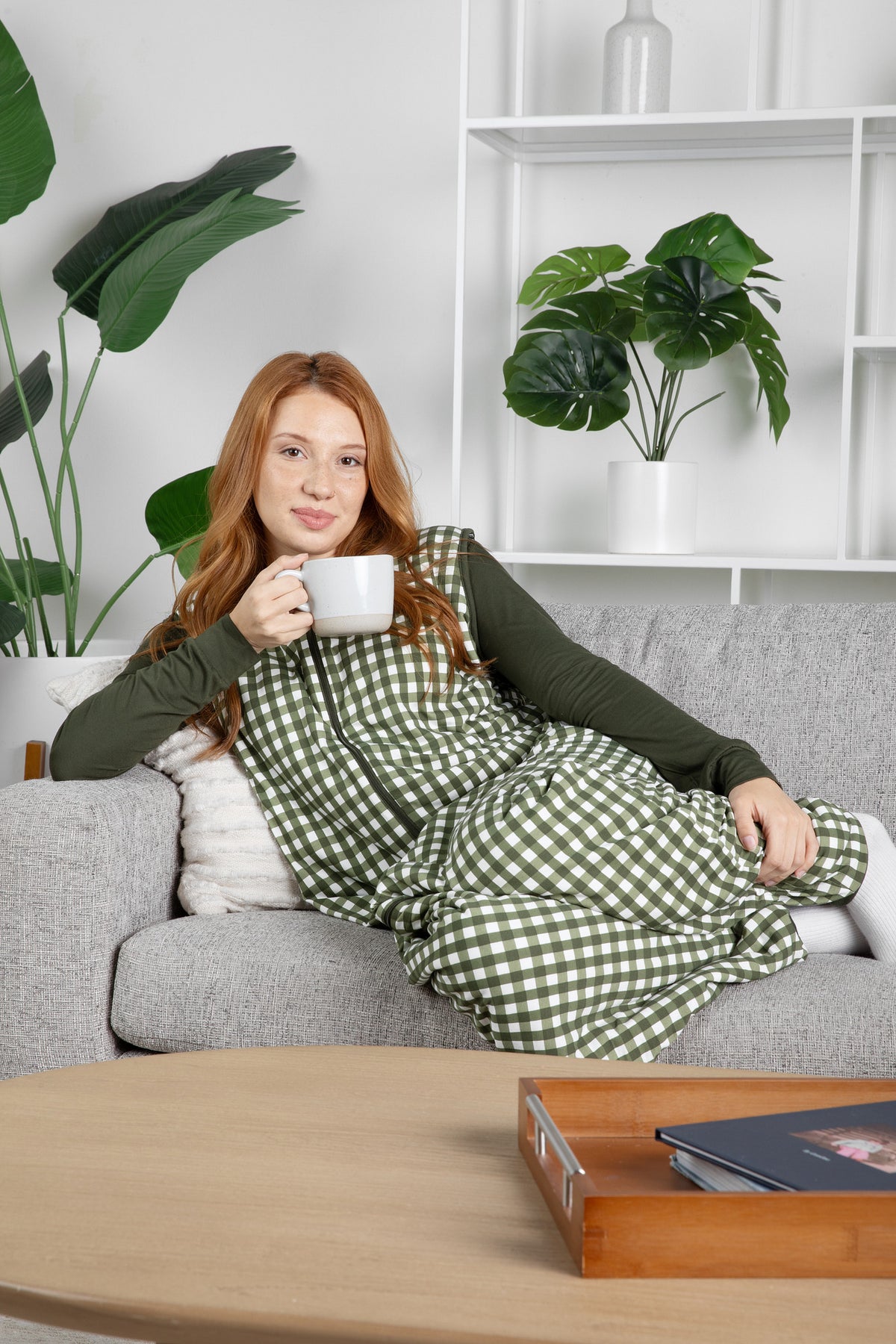 Female modeling the Adult Sleep Bag Walker in Gingham Fir sitting on a couch with a coffee cup wearing the Adult Sleep Bag Walker in Gingham Fir