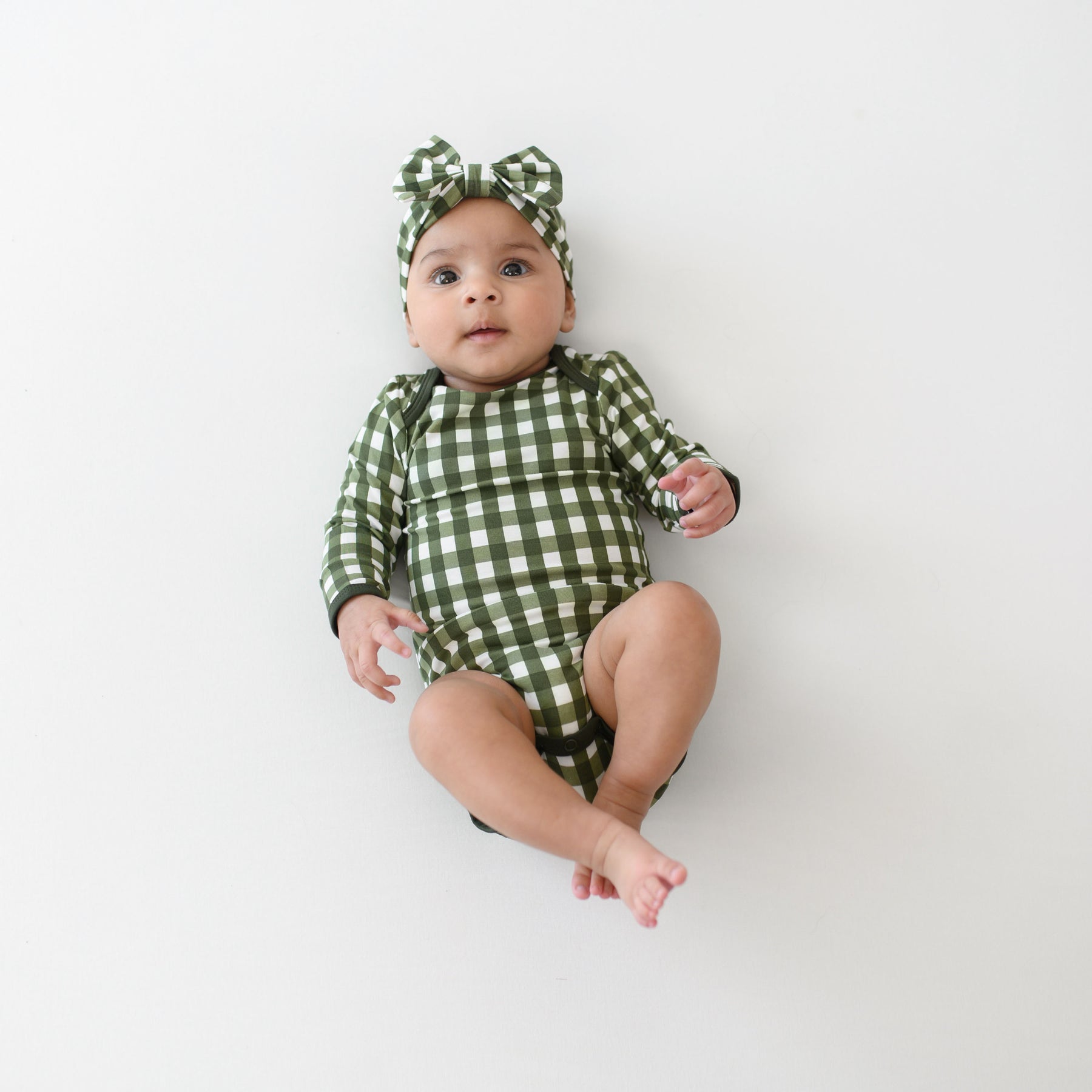 Infant laying down with legs up and crossed at the ankles wearing the Long Sleeve Bodysuit in Gingham Fir with matching bow headband