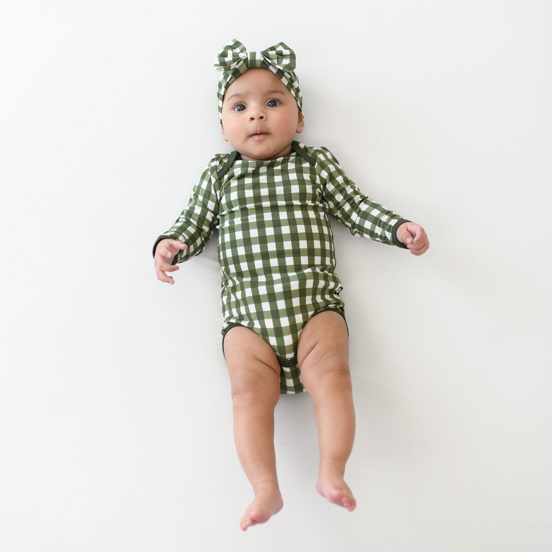 Infant laying down wearing the Long Sleeve Bodysuit in Gingham Fir with matching bow headband