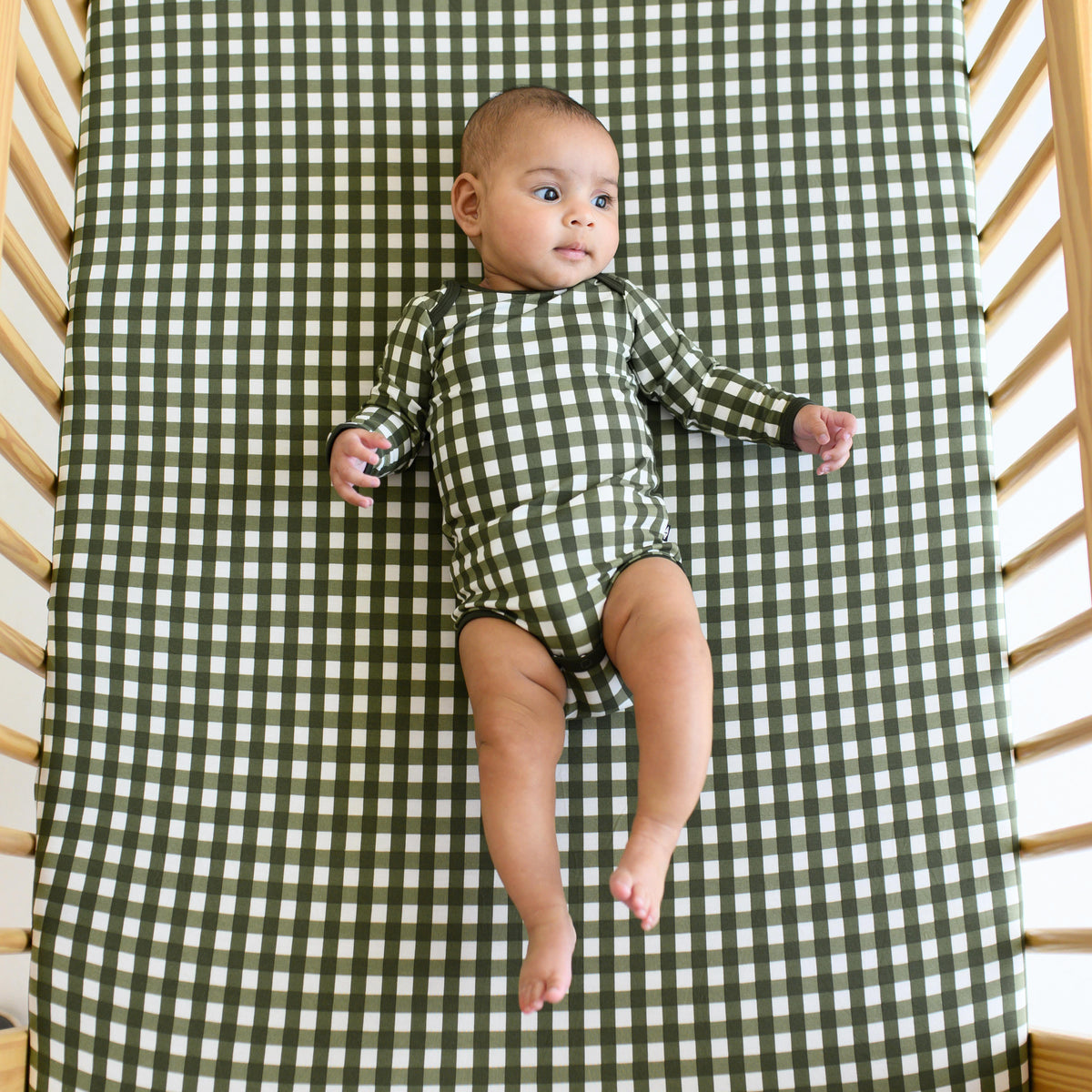 Infant laying in a crib on top of the Crib Sheet in Gingham Fir wearing a matching long sleeve bodysuit