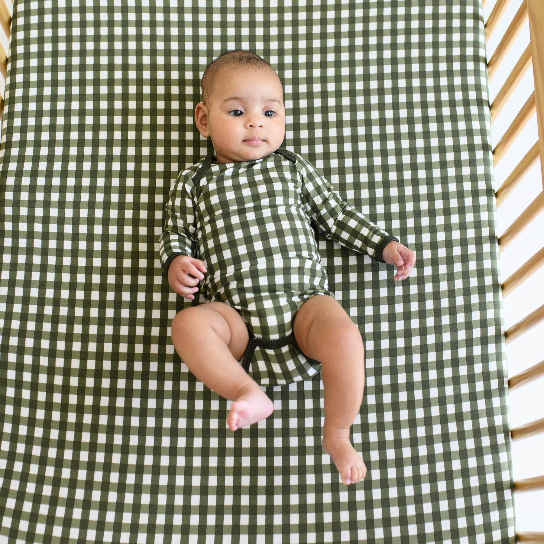 Infant laying in a crib on a Crib Sheet in Gingham Fir wearing the Long Sleeve Bodysuit in Gingham Fir