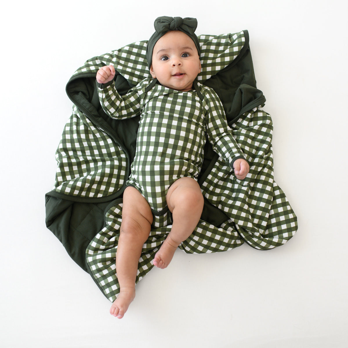 Infant laying on a Kyte Baby Blanket in Gingham Fir wearing a matching long sleeve bodysuit and headband bow
