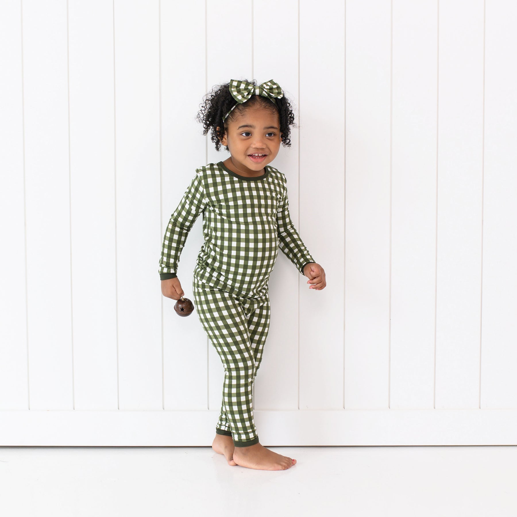 Young girls standing in front of a white paneled wall wearing the Long Sleeve Pajamas in Gingham Fir with matching bow headband