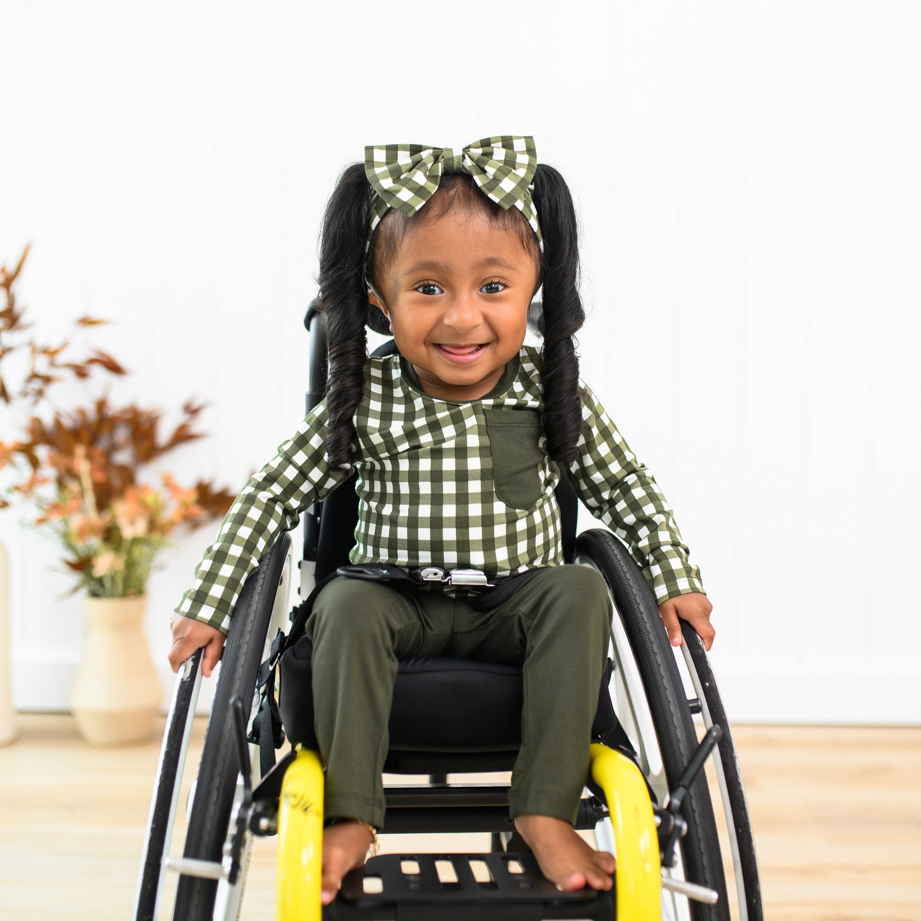 Little girl sitting in her wheelchair wearing the Long Sleeve Toddler Crew Neck Tee in Gingham Fir and matching bow headband