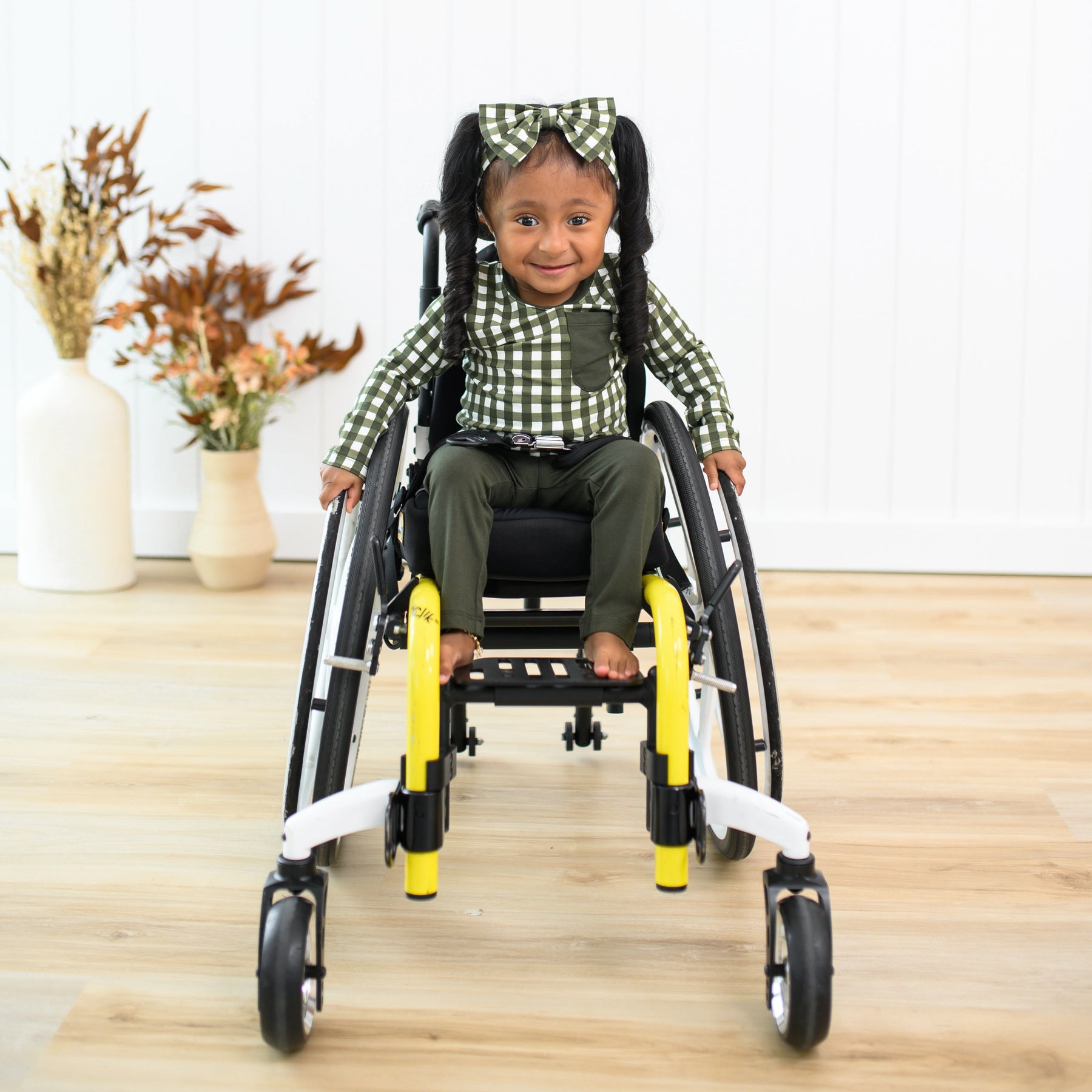 Young girl sitting in her wheelchair wearing the Long Sleeve Toddler Crew Neck Tee in Gingham Fir, Fir leggings and Gingham Fir bow Headband