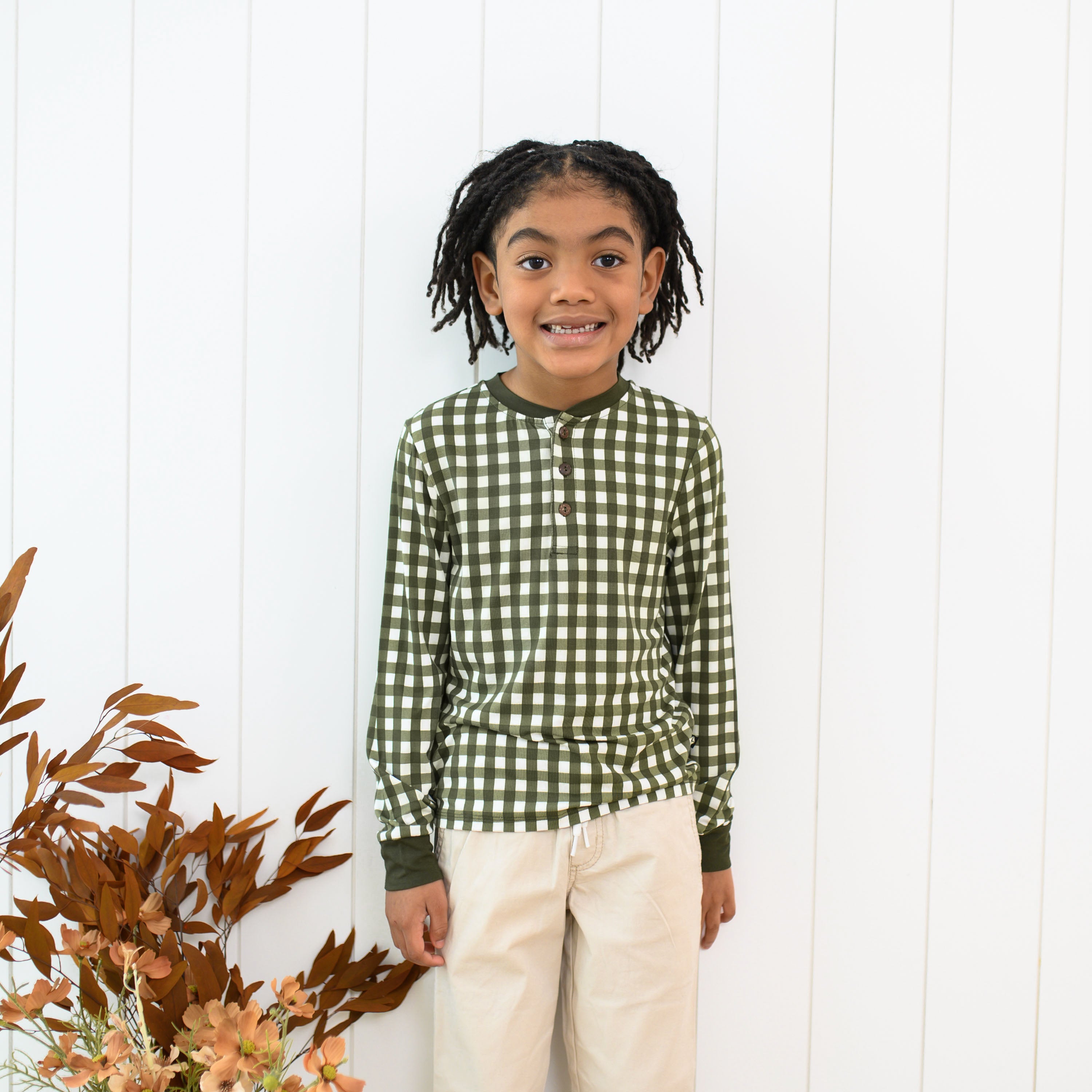 Young girl standing in front of a white paneled wall wearing the Long Sleeve Toddler Henley Tee in Gingham Fir