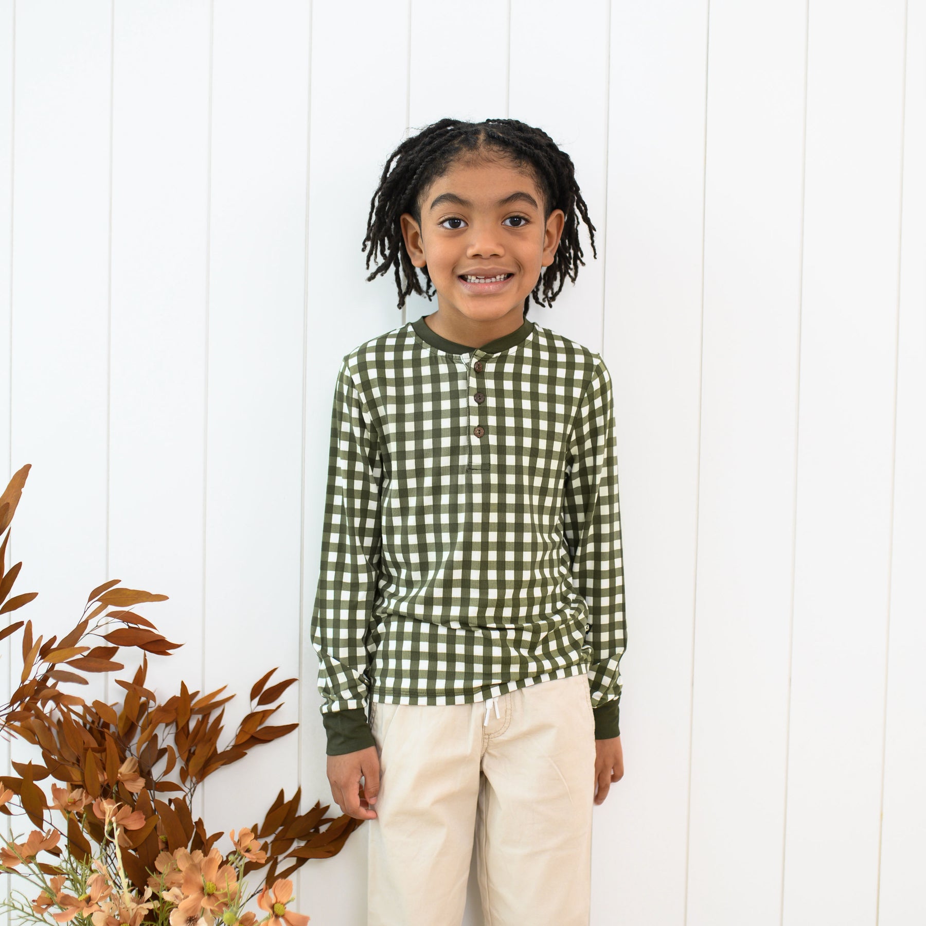 Young girl standing in front of a white paneled wall wearing the Long Sleeve Toddler Henley Tee in Gingham Fir