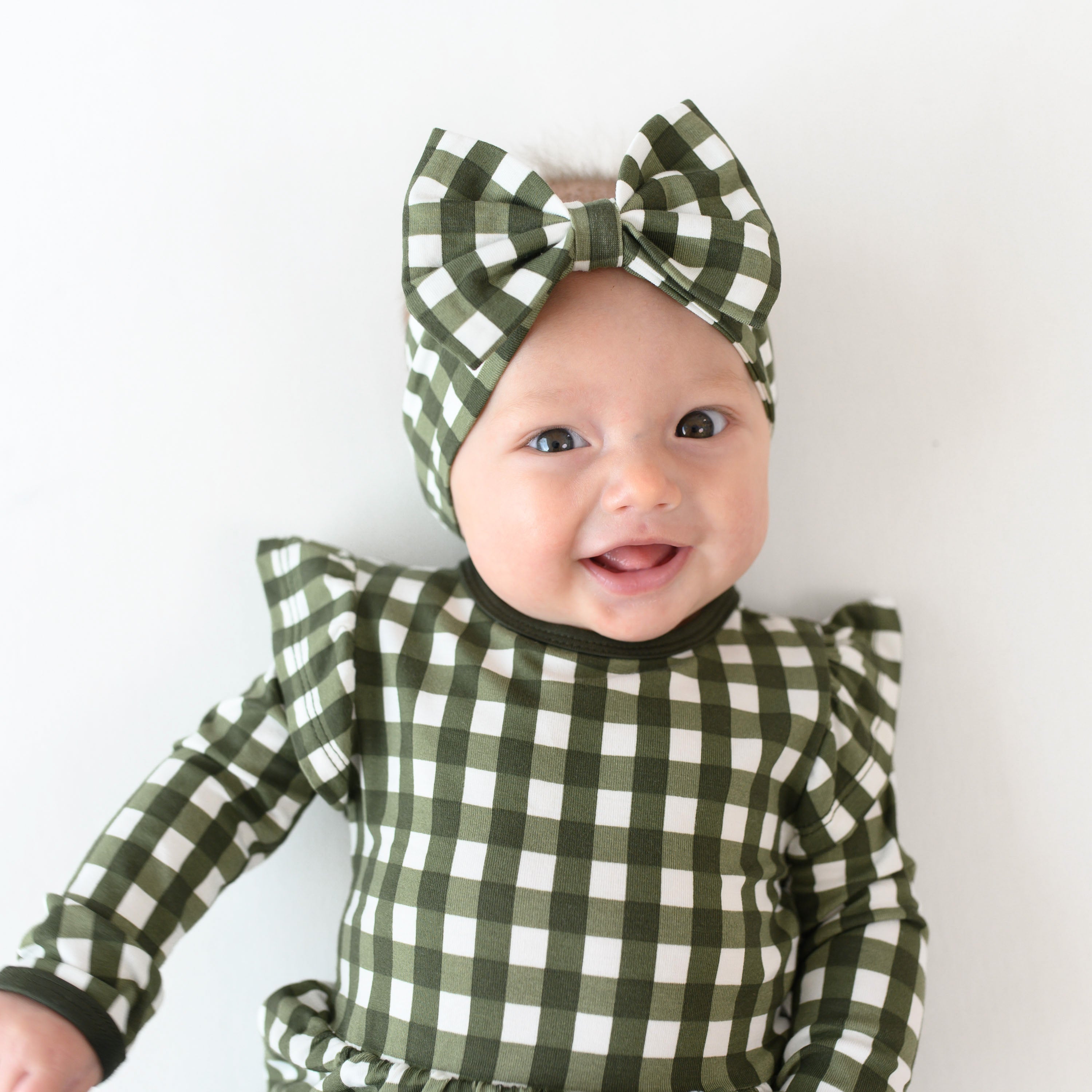 Close up of infant girl wearing the Long Sleeve Twirl Bodysuit Dress in Gingham Fir and matching bow headband