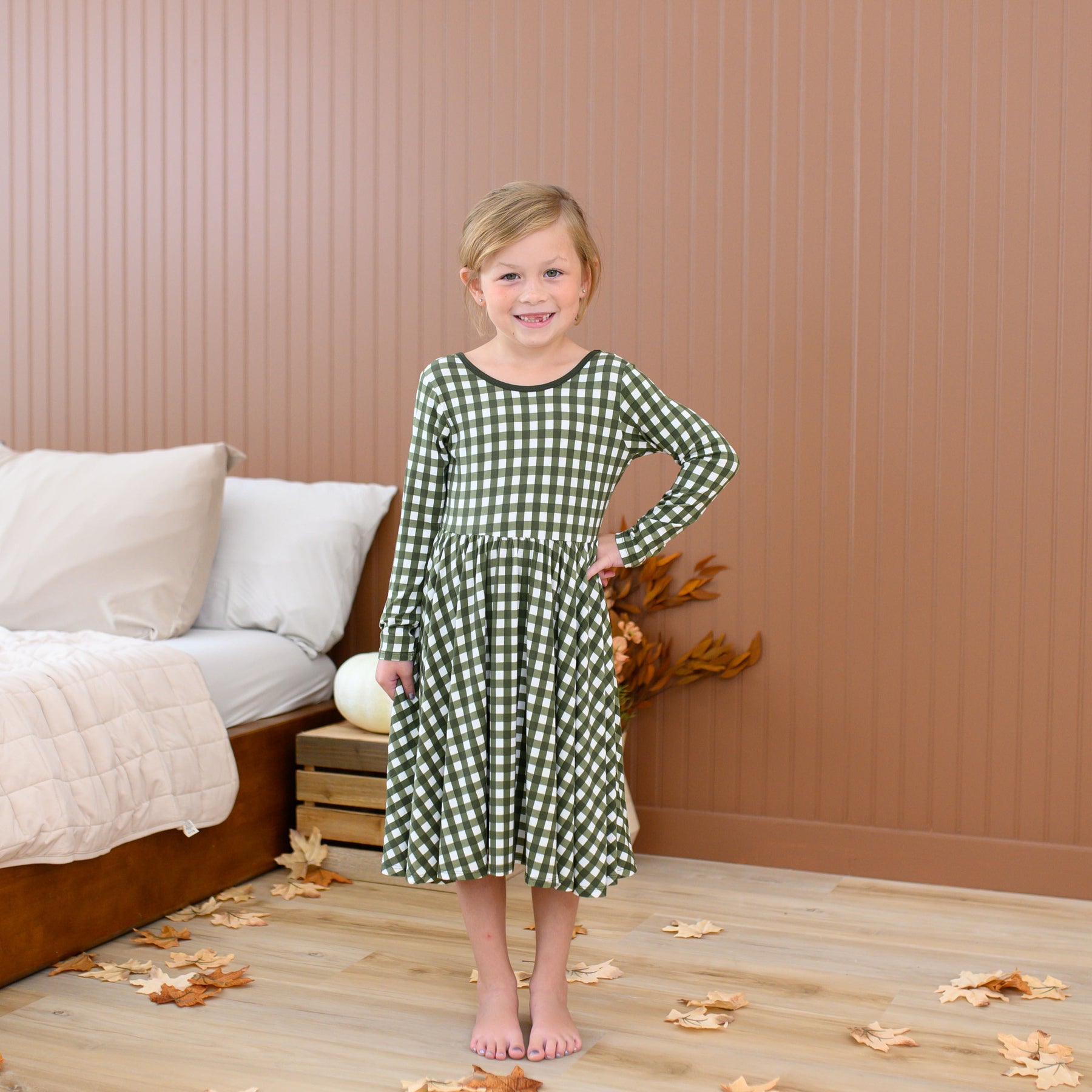Girl standing with one girl on her hip wearing the Long Sleeve Twirl Dress in Gingham Fir standing beside a bed