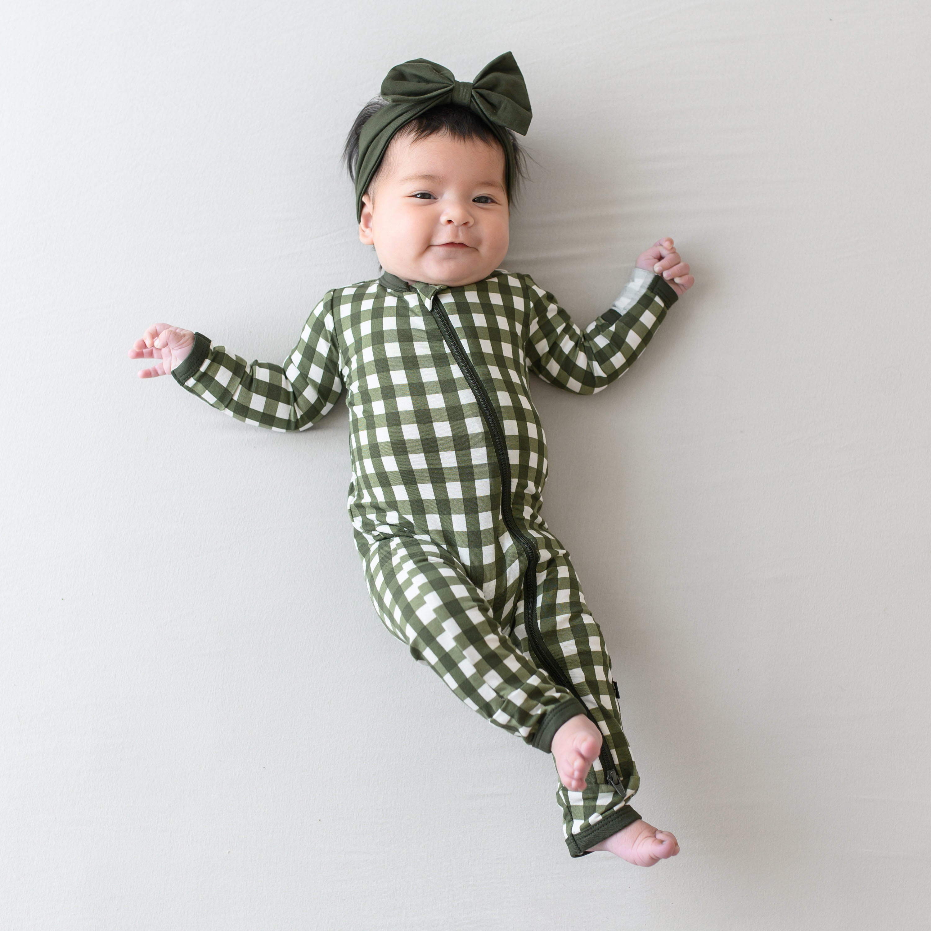 Infant model laying down on a light colored blanket wearing the Zippered Romper in Gingham Fir with a Fir bow Headband