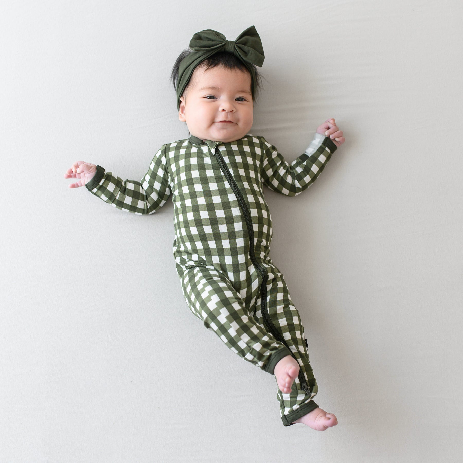 Infant model laying down on a light colored blanket wearing the Zippered Romper in Gingham Fir with a Fir bow Headband