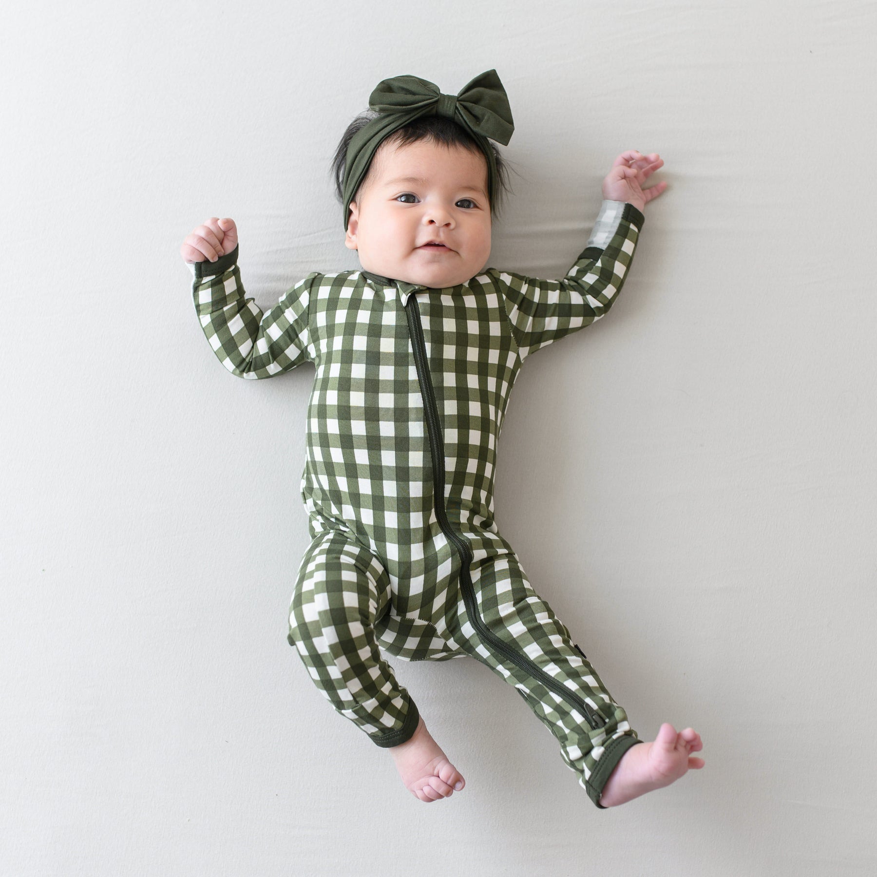 Infant girl laying down wearing the Zippered Romper in Gingham Fir with Fir Bow Headband