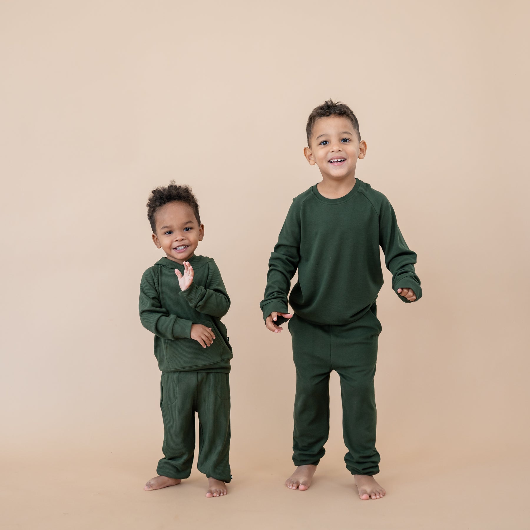 Two young boys matching in Fir in front of a taupe colored backdrop. Youngest boy in the Ribbed Hoodie in Fir with ribbed joggers and old boy is wearing the bamboo jersey jogger set