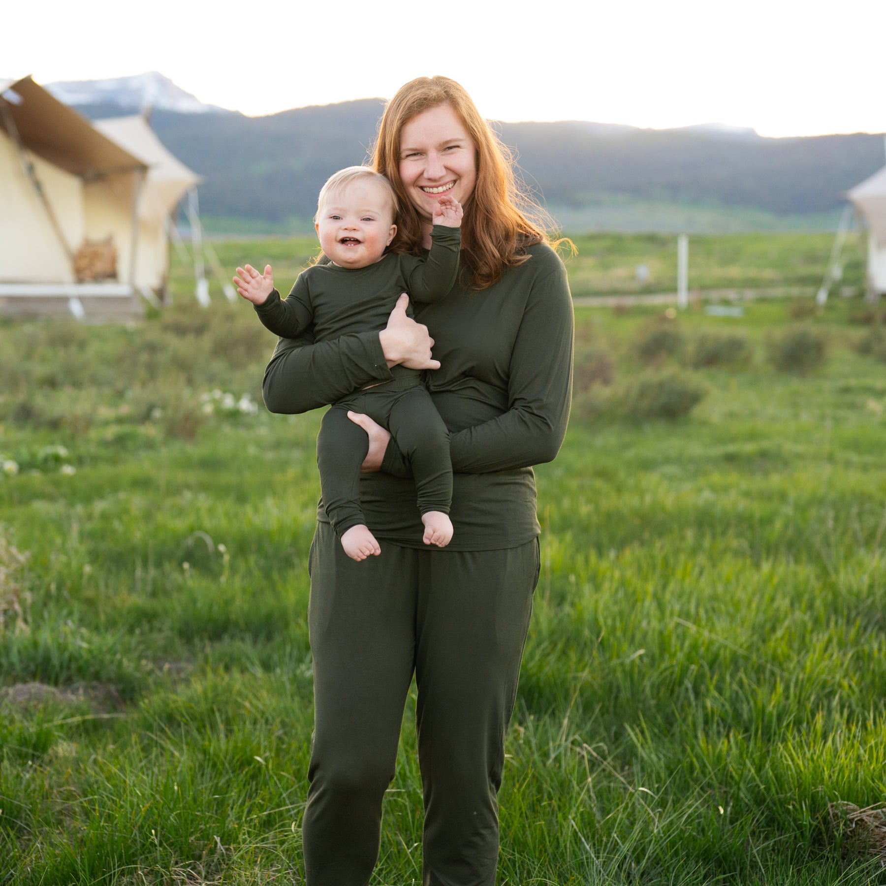 Woman holding a baby in a grassy field with mountains in the background wearing matching pajamas