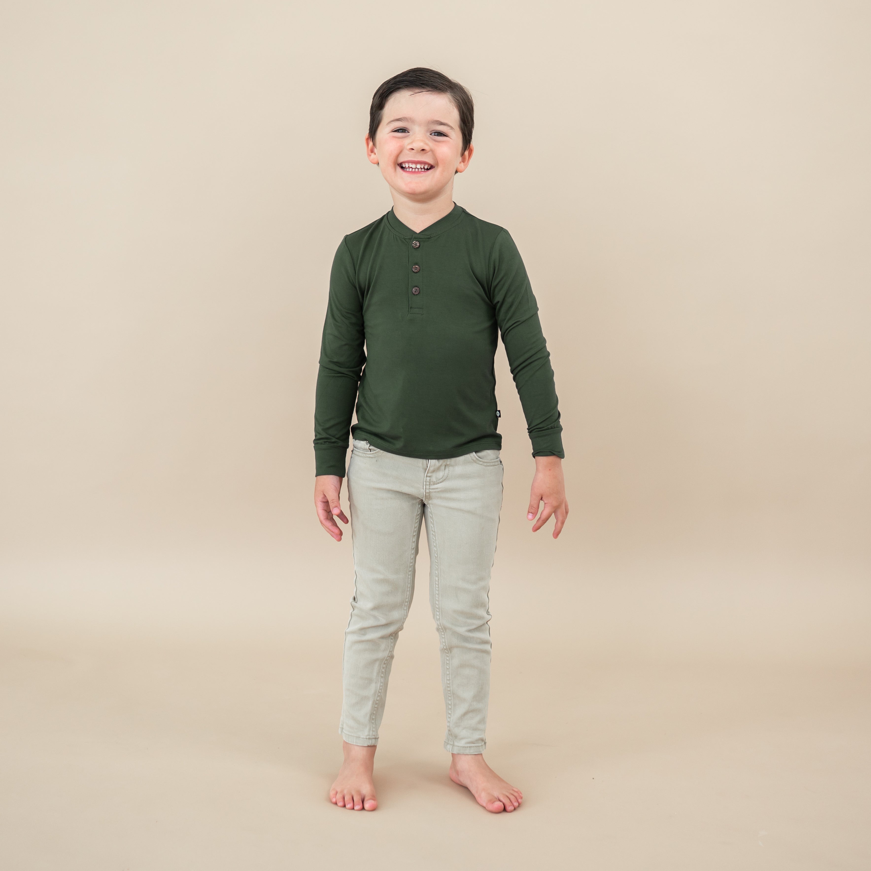 Toddler smiling wearing Long Sleeve Toddler Henley Tee in Fir on tan backdrop