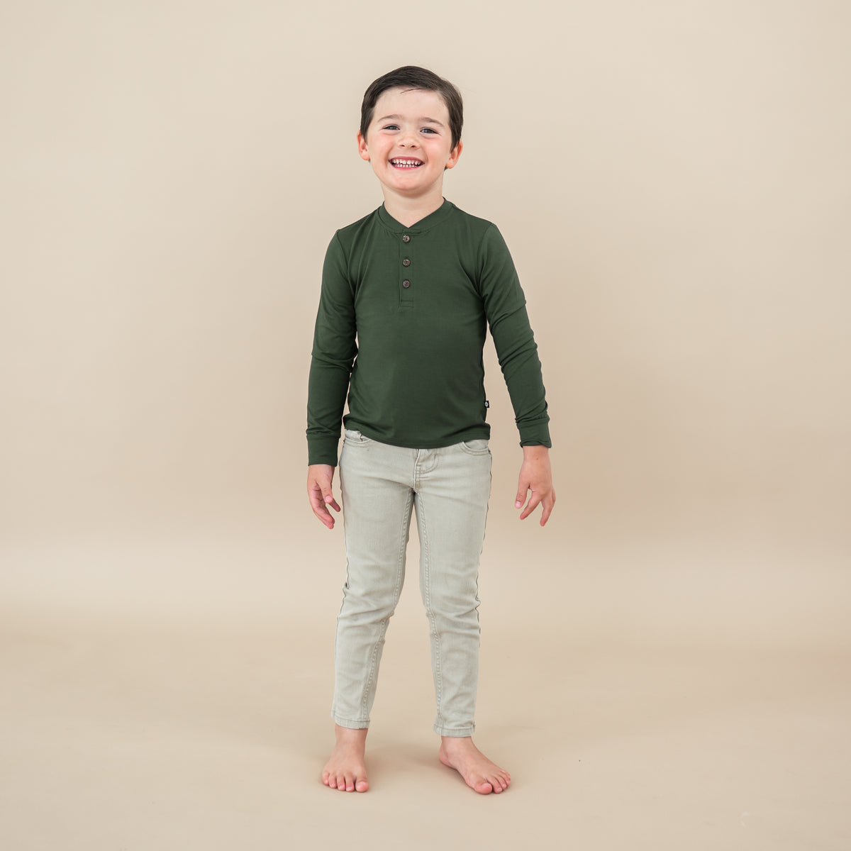Toddler smiling wearing Long Sleeve Toddler Henley Tee in Fir on tan backdrop