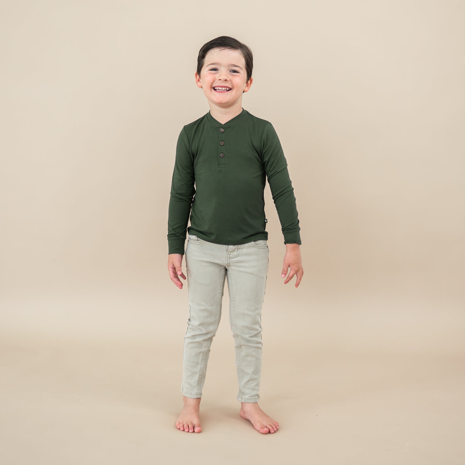 Toddler smiling wearing Long Sleeve Toddler Henley Tee in Fir on tan backdrop
