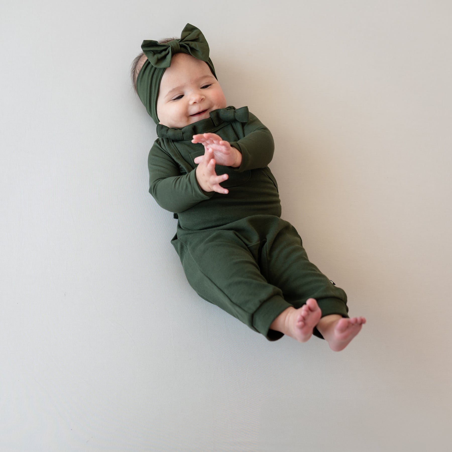Infant girl laying with her feet up in the air wearing the Bamboo Jersey Overall in Fir with matching long sleeve bodysuit on underneath and bow headband as an accessory