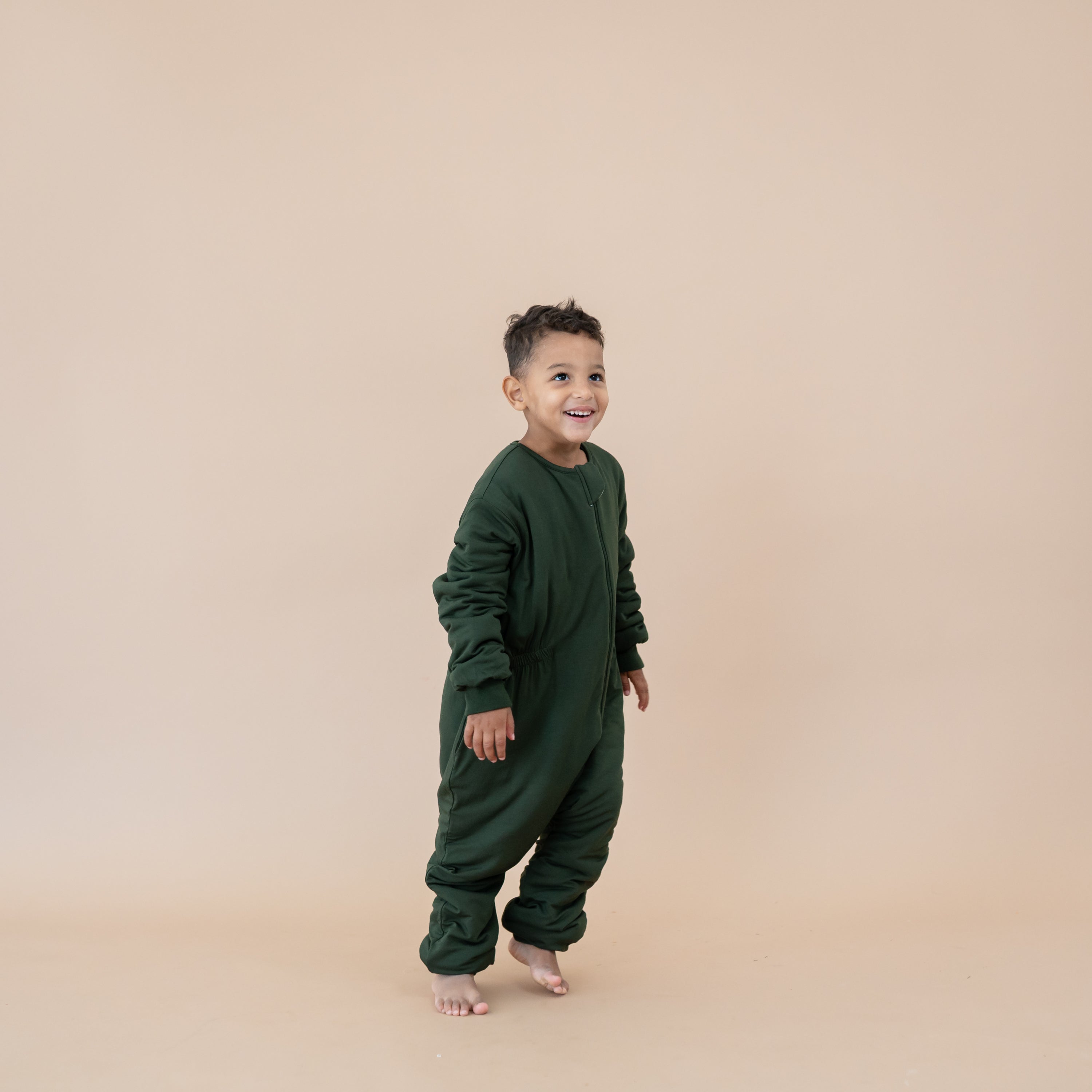 Smiling boy walking wearing the Cozy Playsuit in Fir in front of a light taupe backdrop