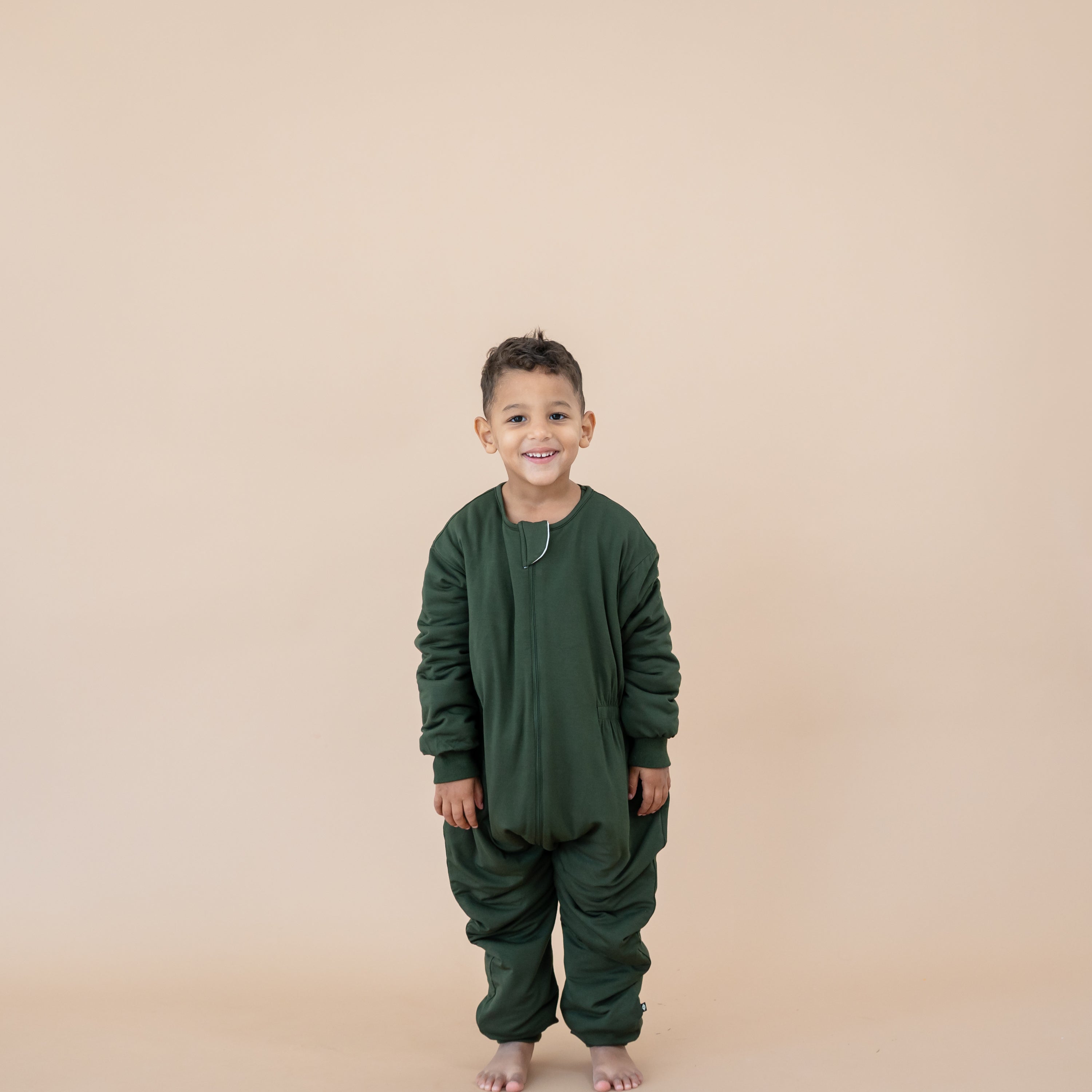 Smiling boy wearing the Cozy Playsuit in Fir in front of a taupe backdrop