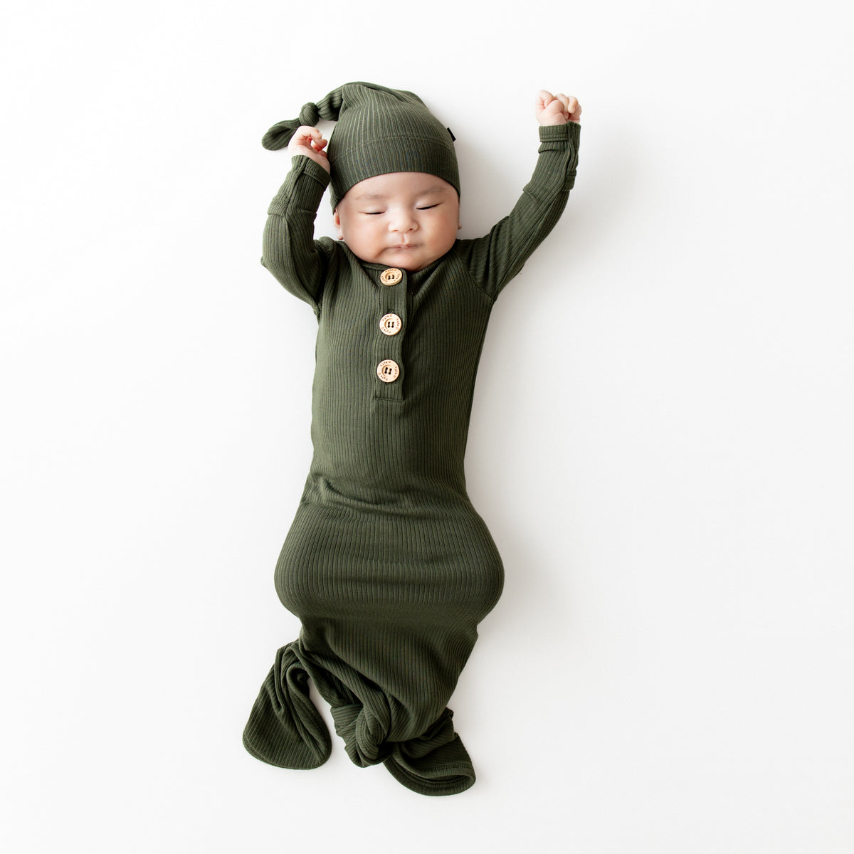 Sleeping infant wearing the Ribbed Knotted Gown with Hat Set in Fir on a light colored backdrop