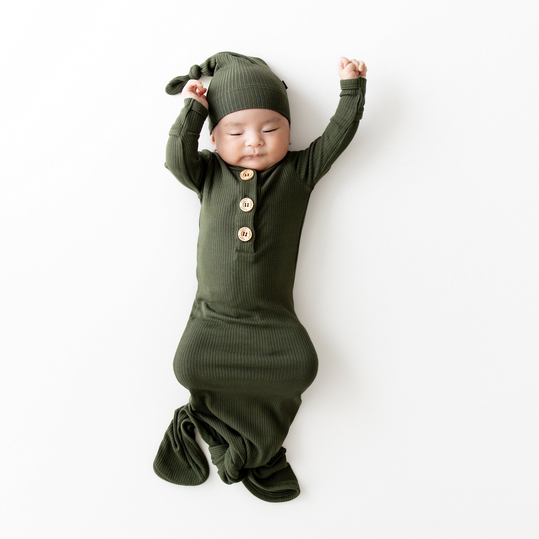 Sleeping infant wearing the Ribbed Knotted Gown with Hat Set in Fir on a light colored backdrop