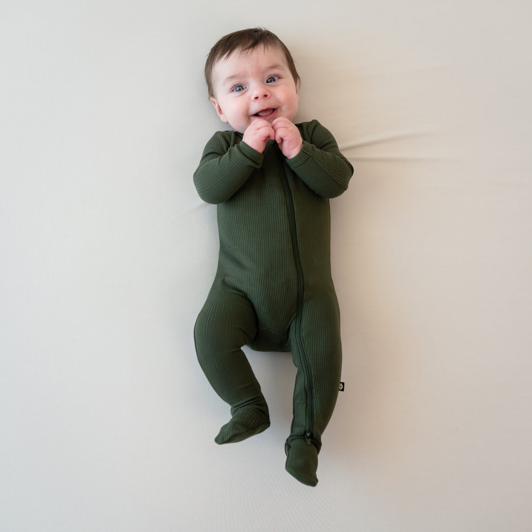 Smiling infant with hands by their mouth wearing the Ribbed Zipper Footie in Fir on a Bisque blanket