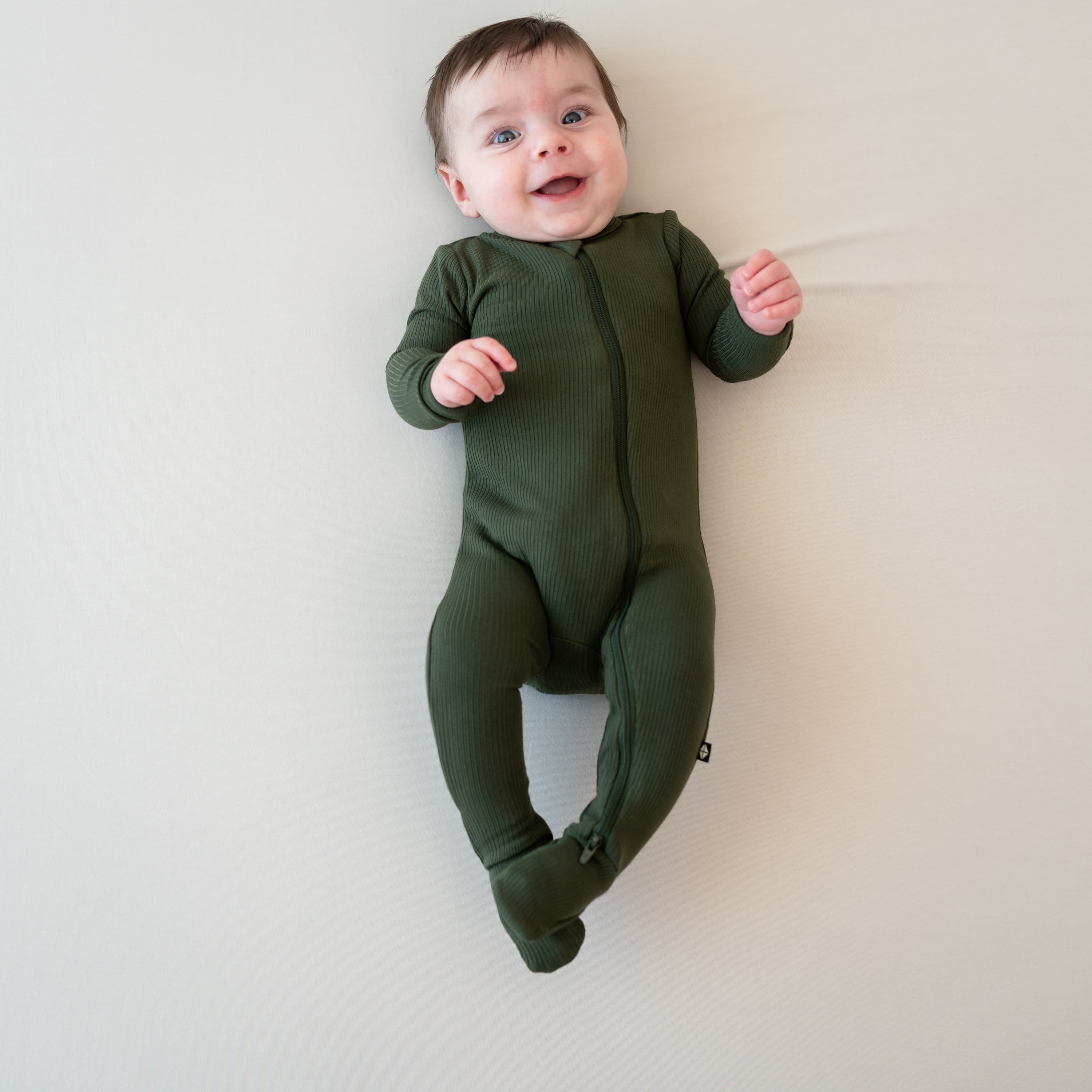 Smiling infant laying on a Bisque blanket wearing the Ribbed Zipper Footie in Fir