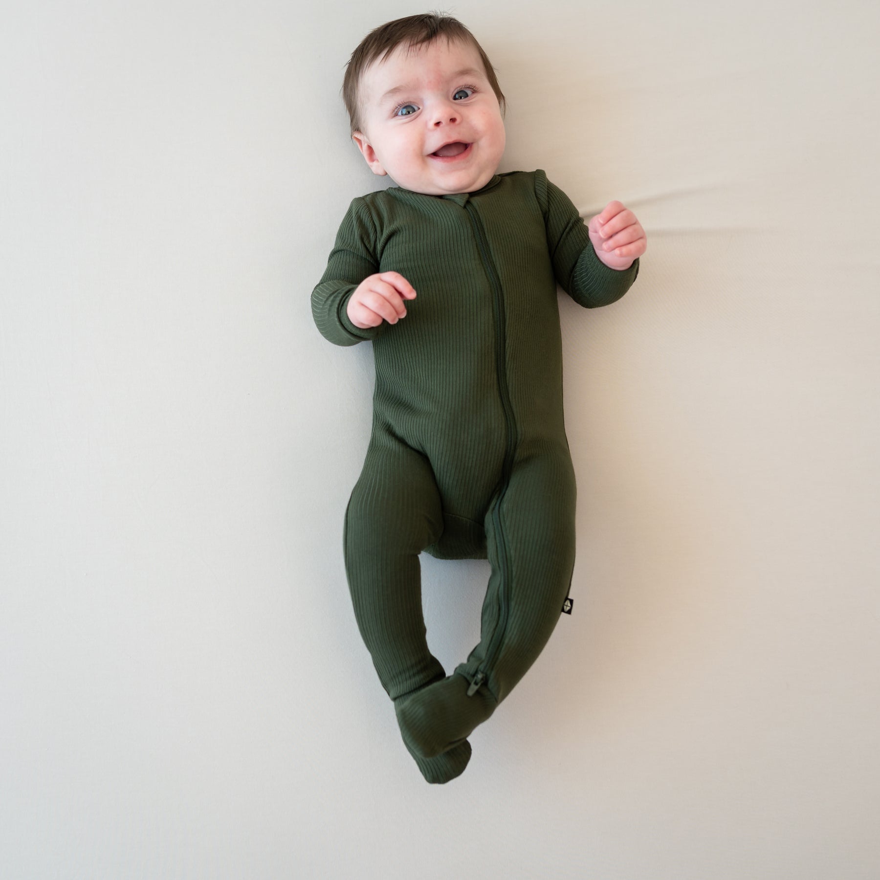 Smiling infant laying on a Bisque blanket wearing the Ribbed Zipper Footie in Fir