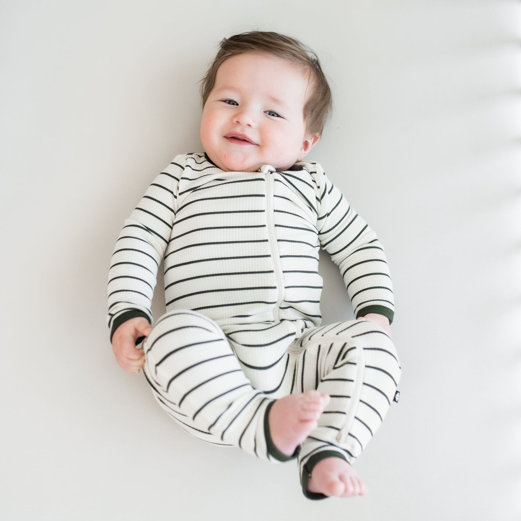 Infant laying down on a light neutral surface wearing the Ribbed Zipper Romper in Fir Stripe with their legs up in the air