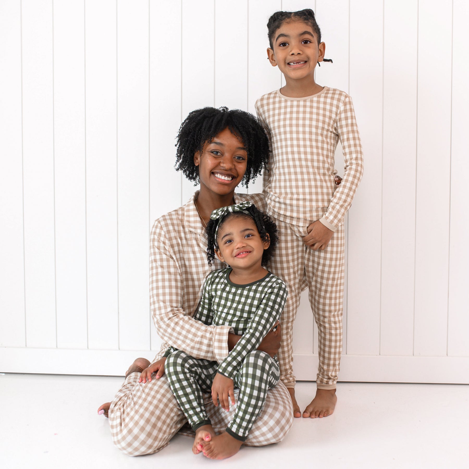 Smiling family matching in Gingham. mother wearing the Long sleeve women's pajama set in Bisque Gingham matching her oldest daughter wearing long sleeve toddler pajamas. Youngest daughter is wearing the Long Sleeve Pajamas in Fir Bisque