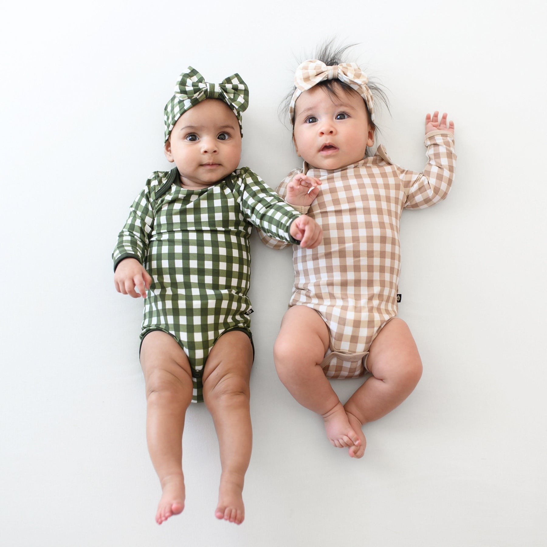 Two infants laying side by side wearing the Long Sleeve Bodysuit in Gingham Fir and Gingham Bisque with matching bow headbands