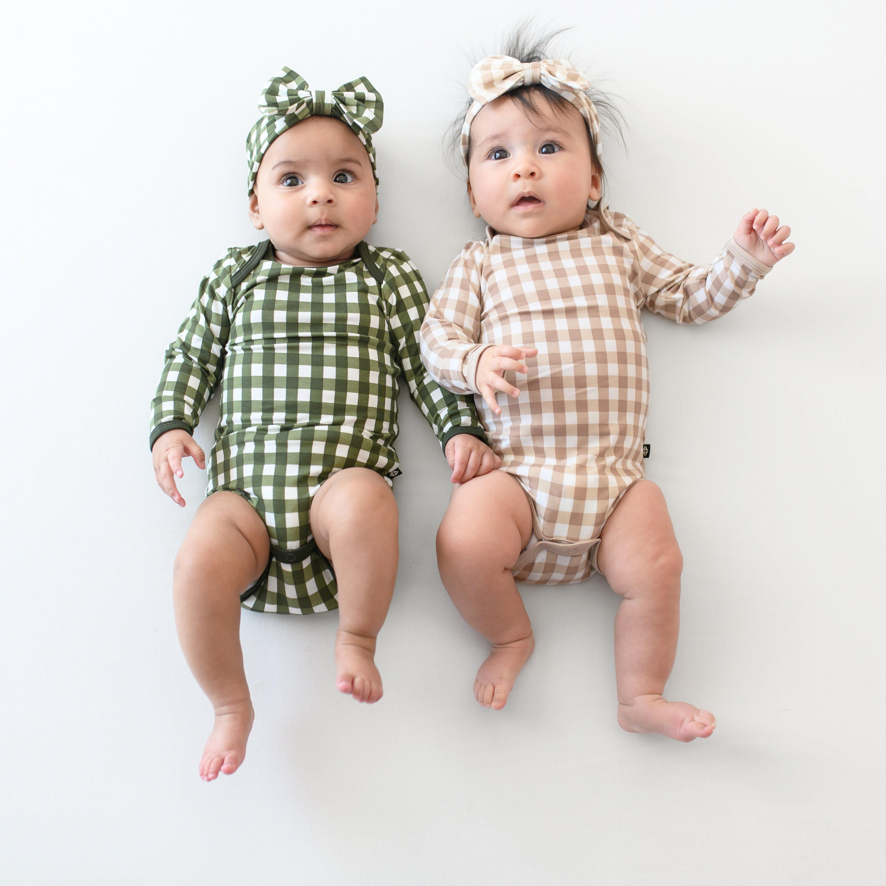Two infant models laying side by side wearing the Long Sleeve Bodysuit in Gingham Bisque and Long Sleeve Bodysuit in Fir Bisque with matching bow headbands