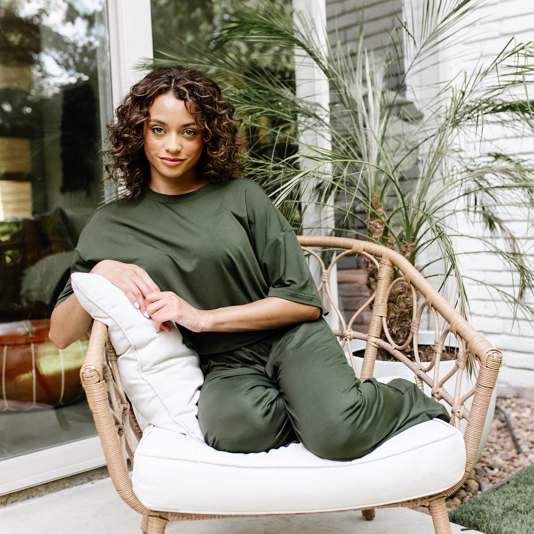 Female model sitting in a wicker chair with white cushions wearing the Women's Short Sleeve Tee in Fir with matching wide leg pants