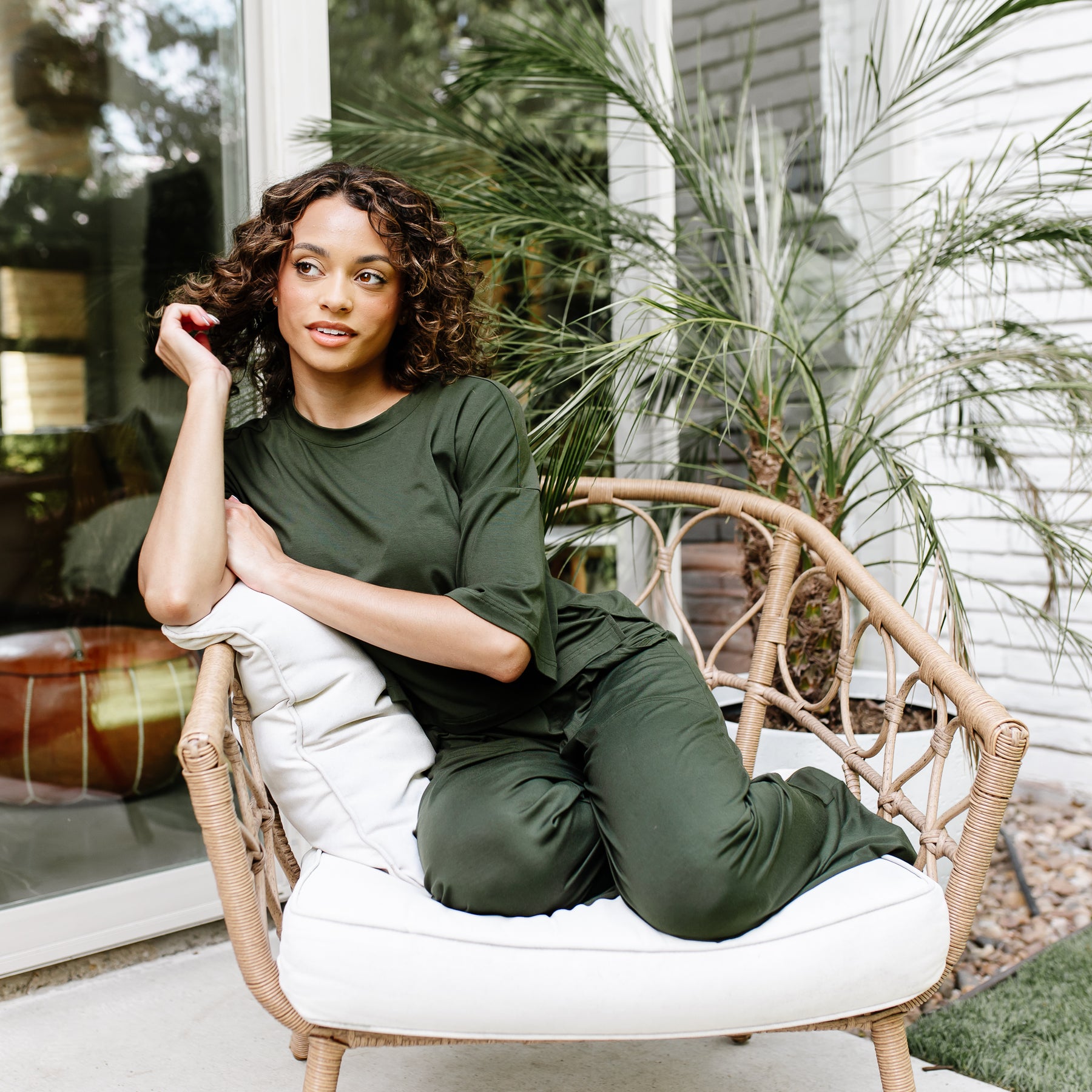 Female model sitting in a wicker chair with white cushions wearing the Women's Wide Leg Pant in Fir with matching short sleeve tee