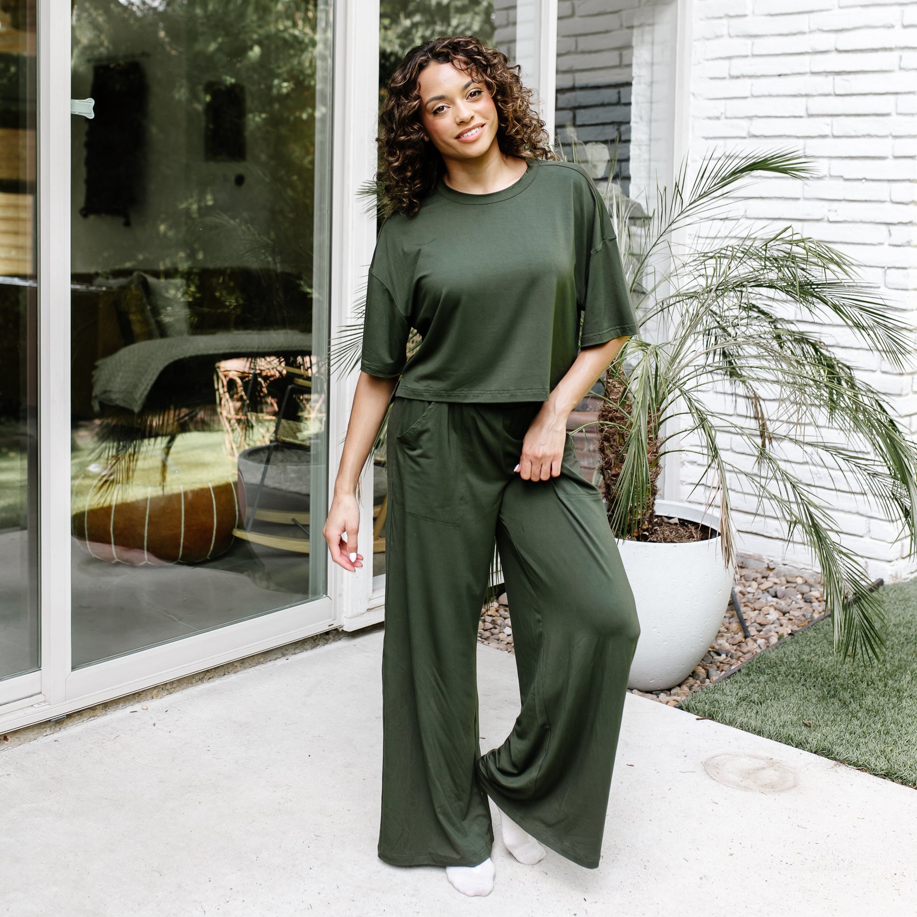 Female model standing in front of glass windows on a white brick house wearing the Women's Short Sleeve Tee in Fir with matching wide leg pants