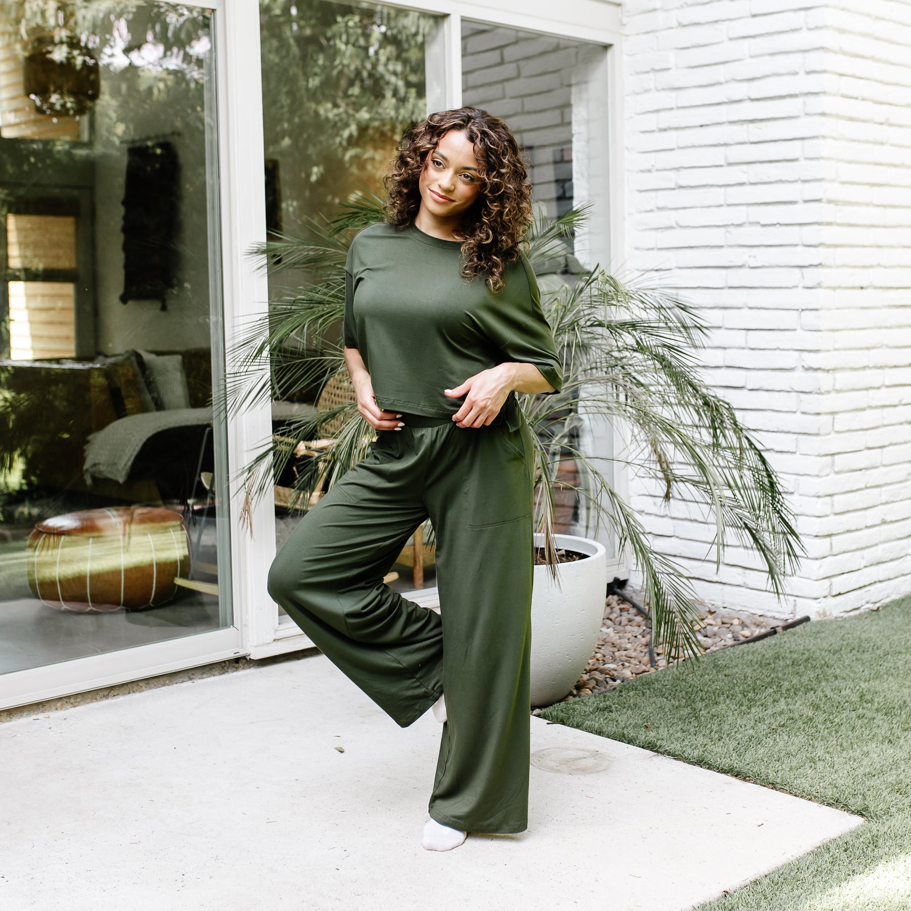 Female standing on a concrete patio in front of glass windows on a white brick house wearing the Women's Wide Leg Pant in Fir with matching short sleeve tee