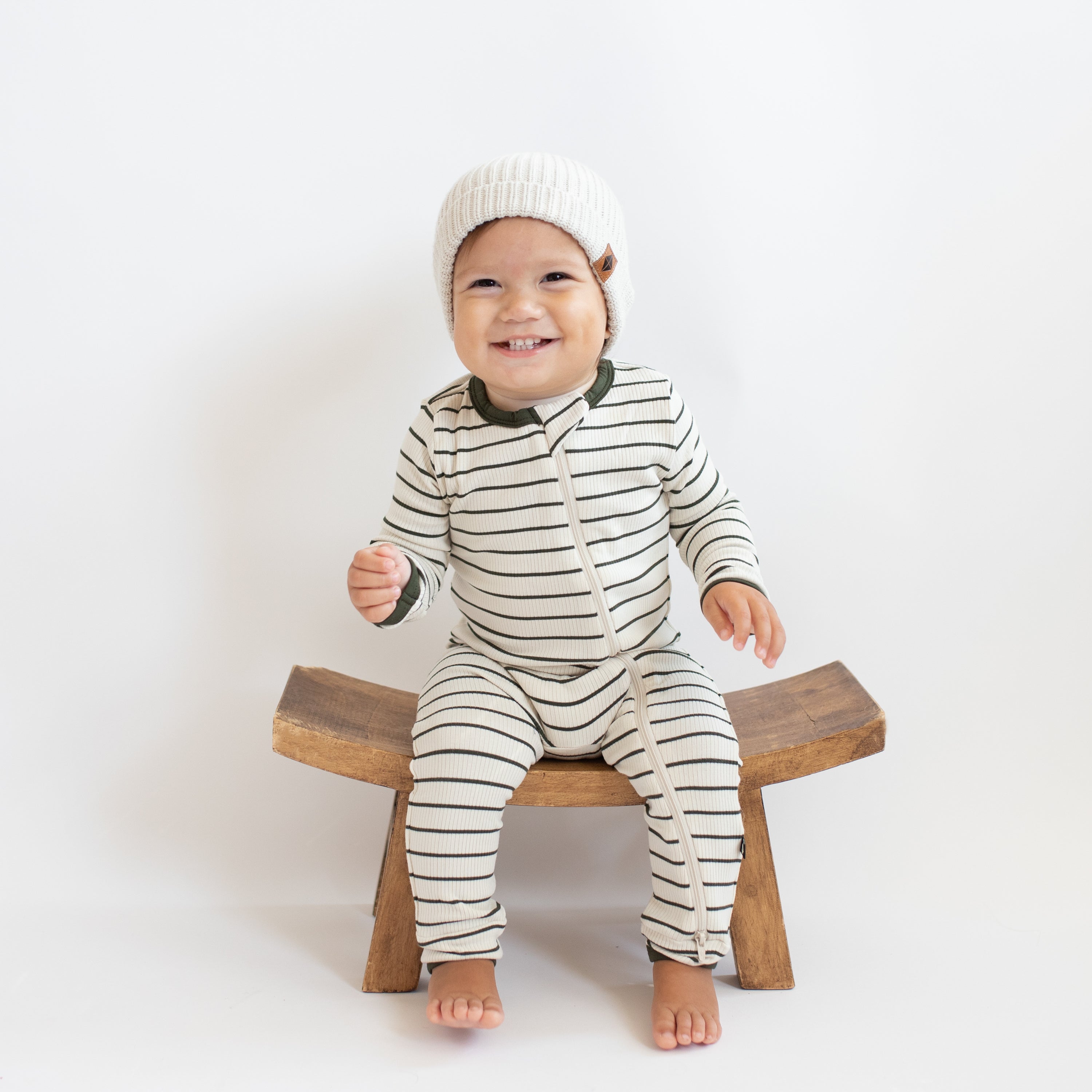 Smiling toddler sitting on a wooden stool wearing the Ribbed Zipper Romper in Fir Stripe and cream colored chunky knit beanie
