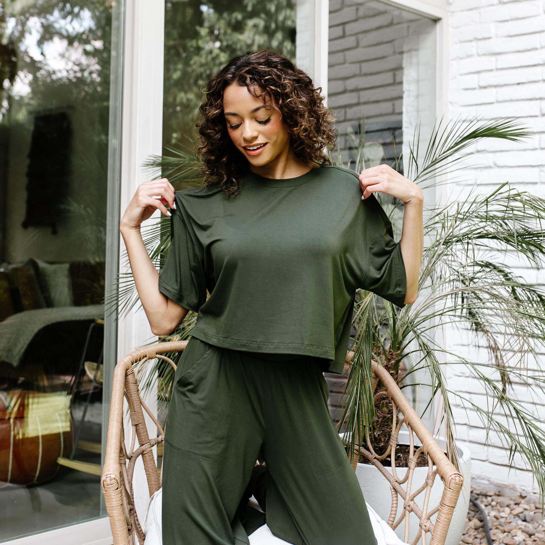 Female model standing in front of a white brick house wearing the Women's Short Sleeve Tee in Fir