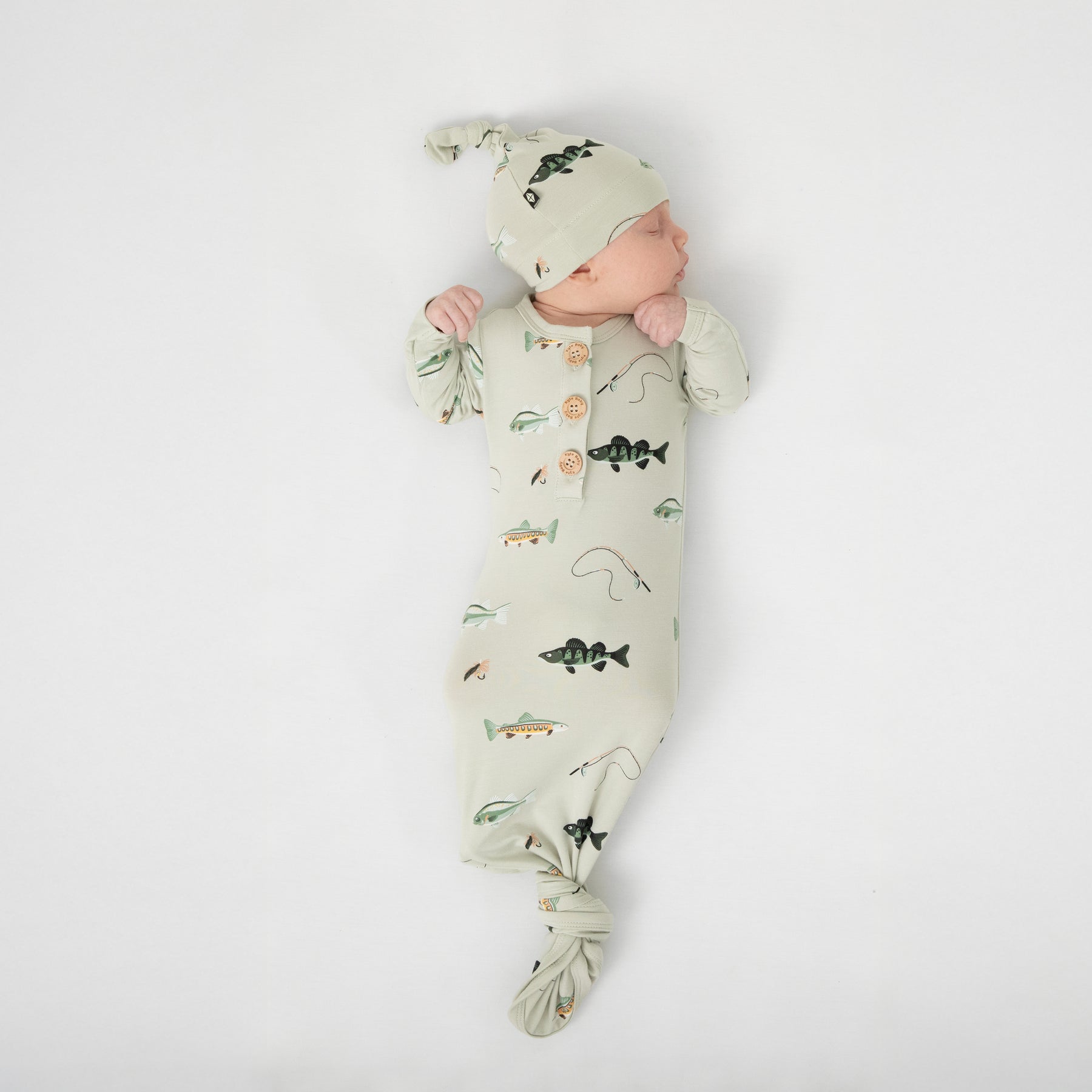 Sleeping newborn laying on a white blanket wearing the Knotted Gown with Hat Set in Fishing tied at the botton